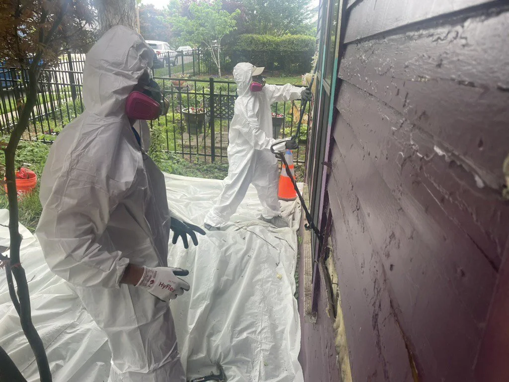 Workers in white protective suits and respirators spray a house exterior while standing on protective plastic sheeting.