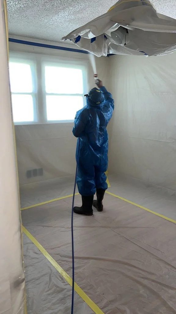 Worker in blue protective coveralls and respirator spraying inside a plastic-lined paint booth