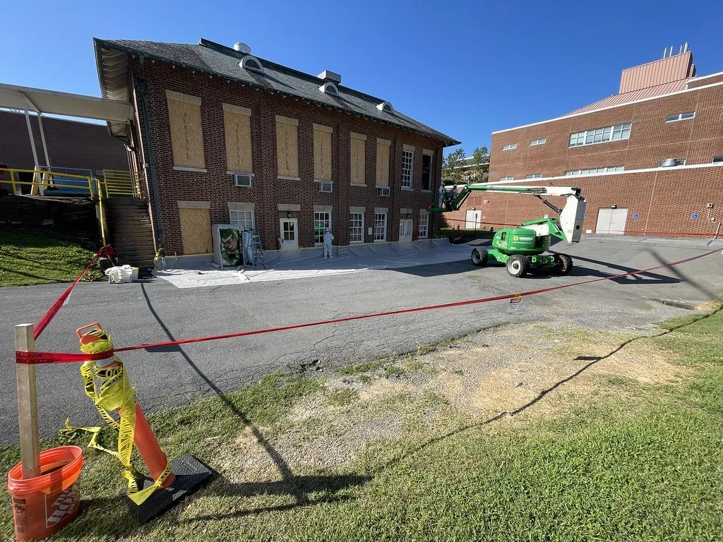 Brick building undergoing window renovation with boarded windows, protective sheeting, and a green boom lift behind caution tape.