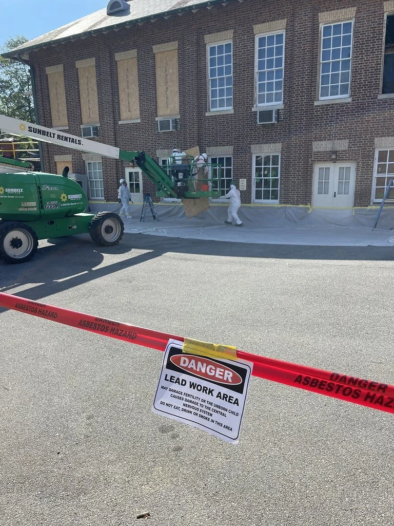 Lead paint and asbestos hazard work area outside a brick building, with workers in protective suits and a green Sunbelt lift.