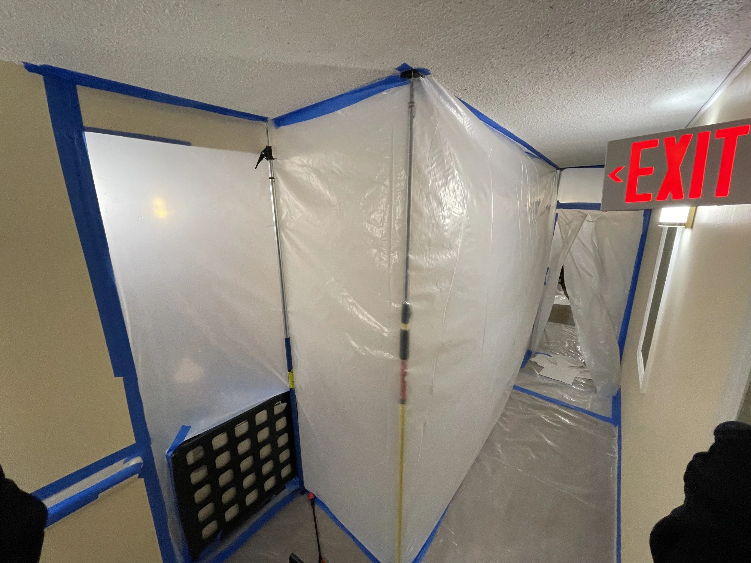 Interior renovation area sealed with plastic sheeting and blue painter’s tape, with an illuminated red EXIT sign.