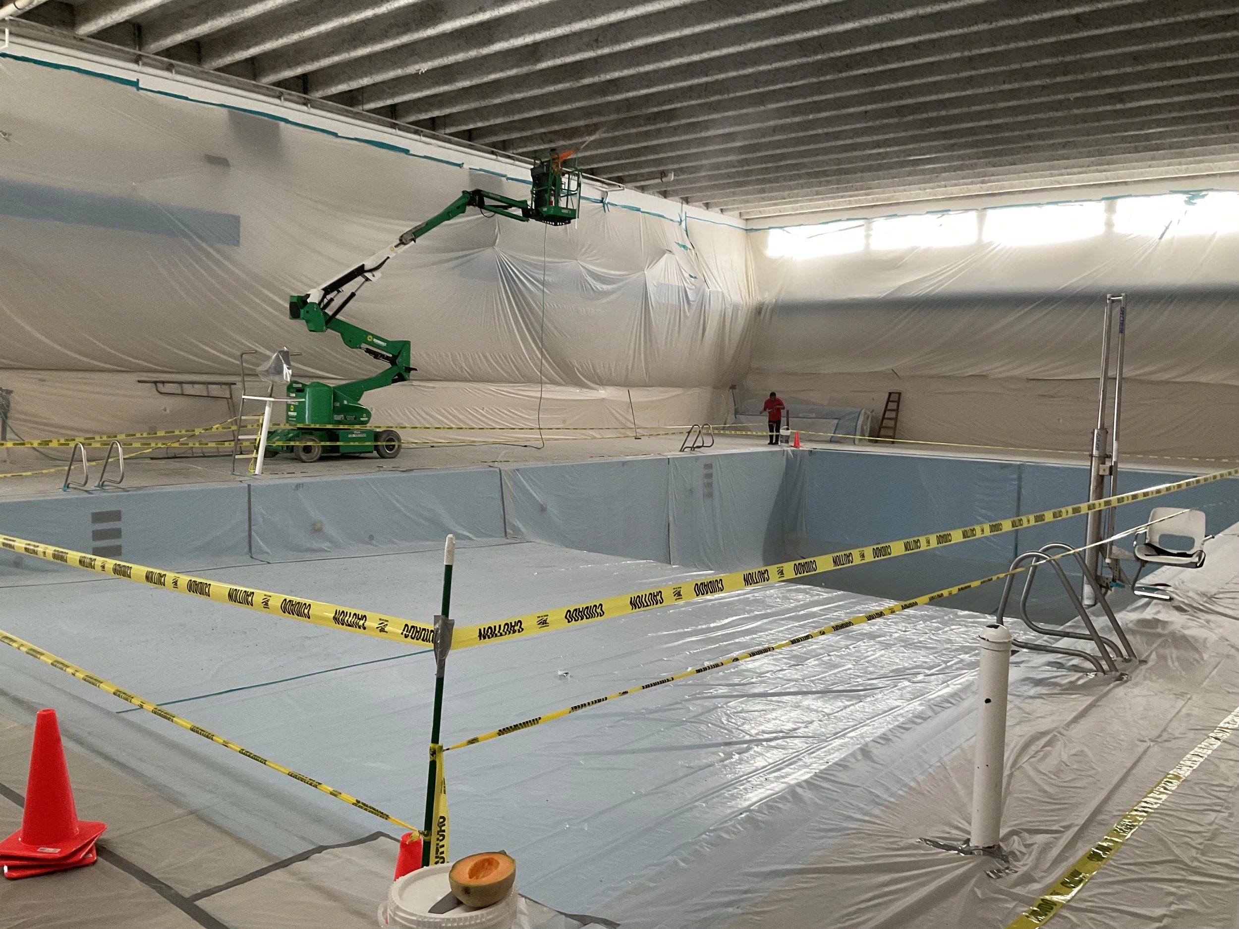 Indoor swimming pool renovation with plastic sheeting, yellow caution tape, and a green boom lift.