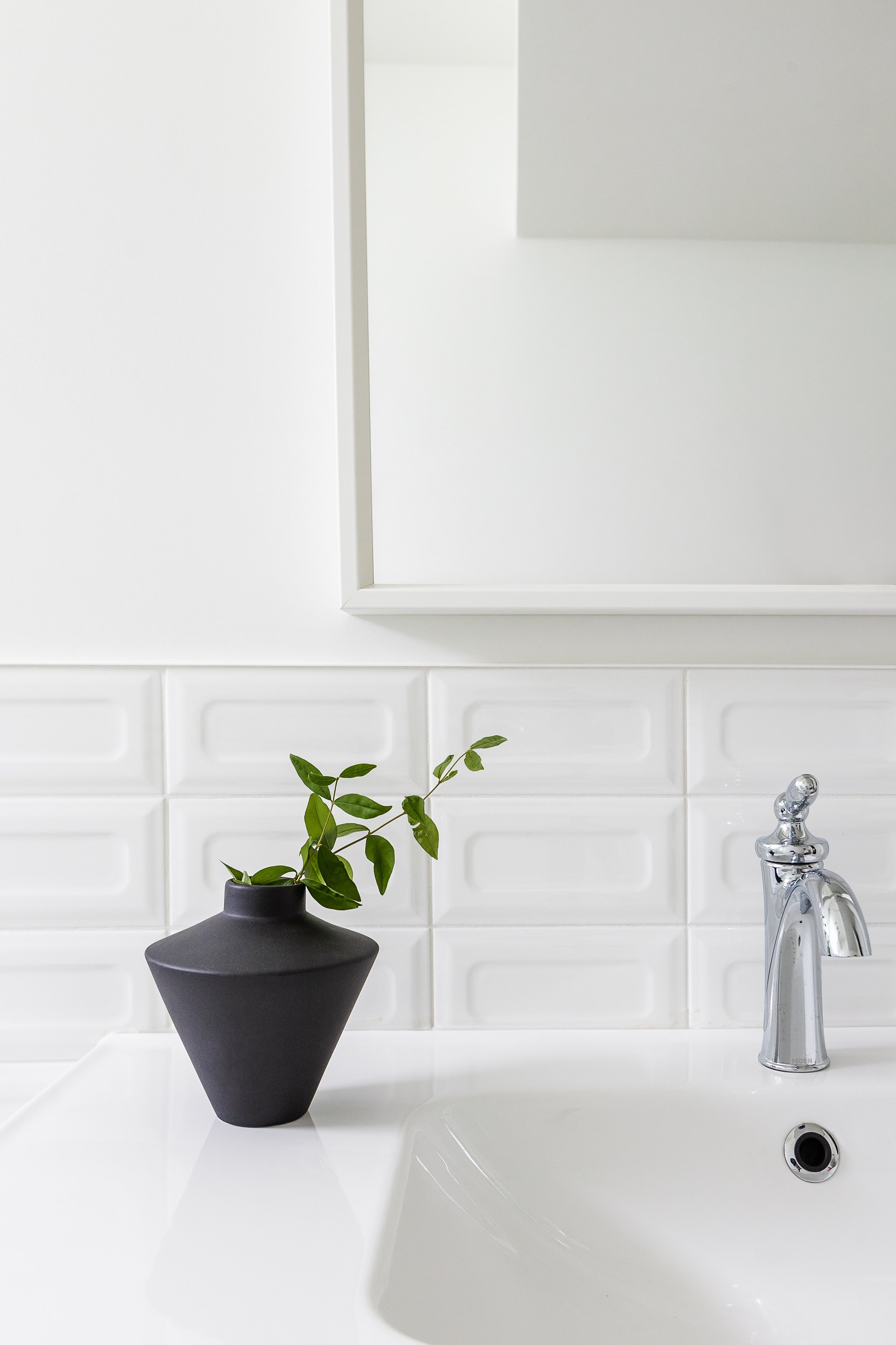 Modern bathroom sink with chrome faucet, black vase with green leaves, and white textured tiles.