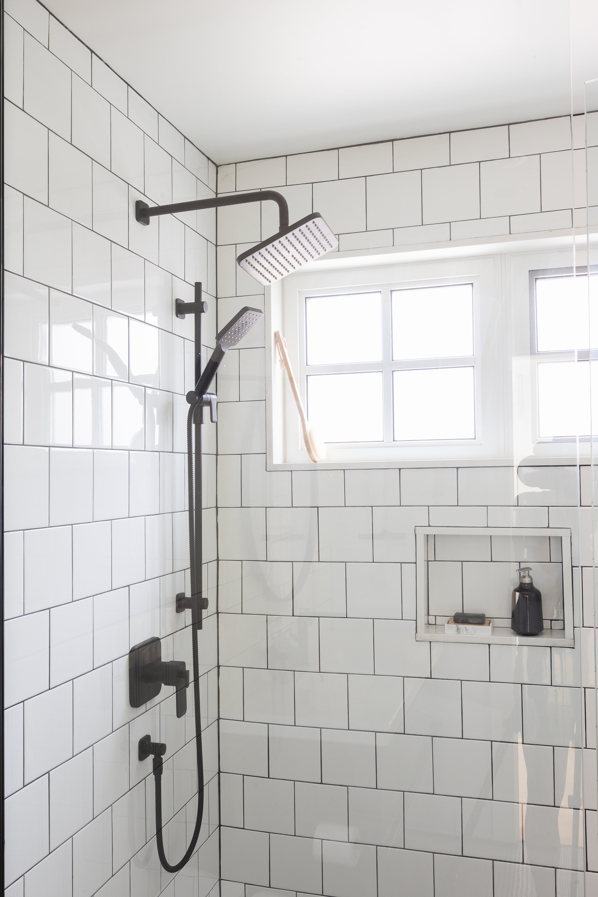 Modern bathroom shower with white subway tiles, square overhead showerhead, handheld shower, window, and built-in shelf holding soap and a bottle.