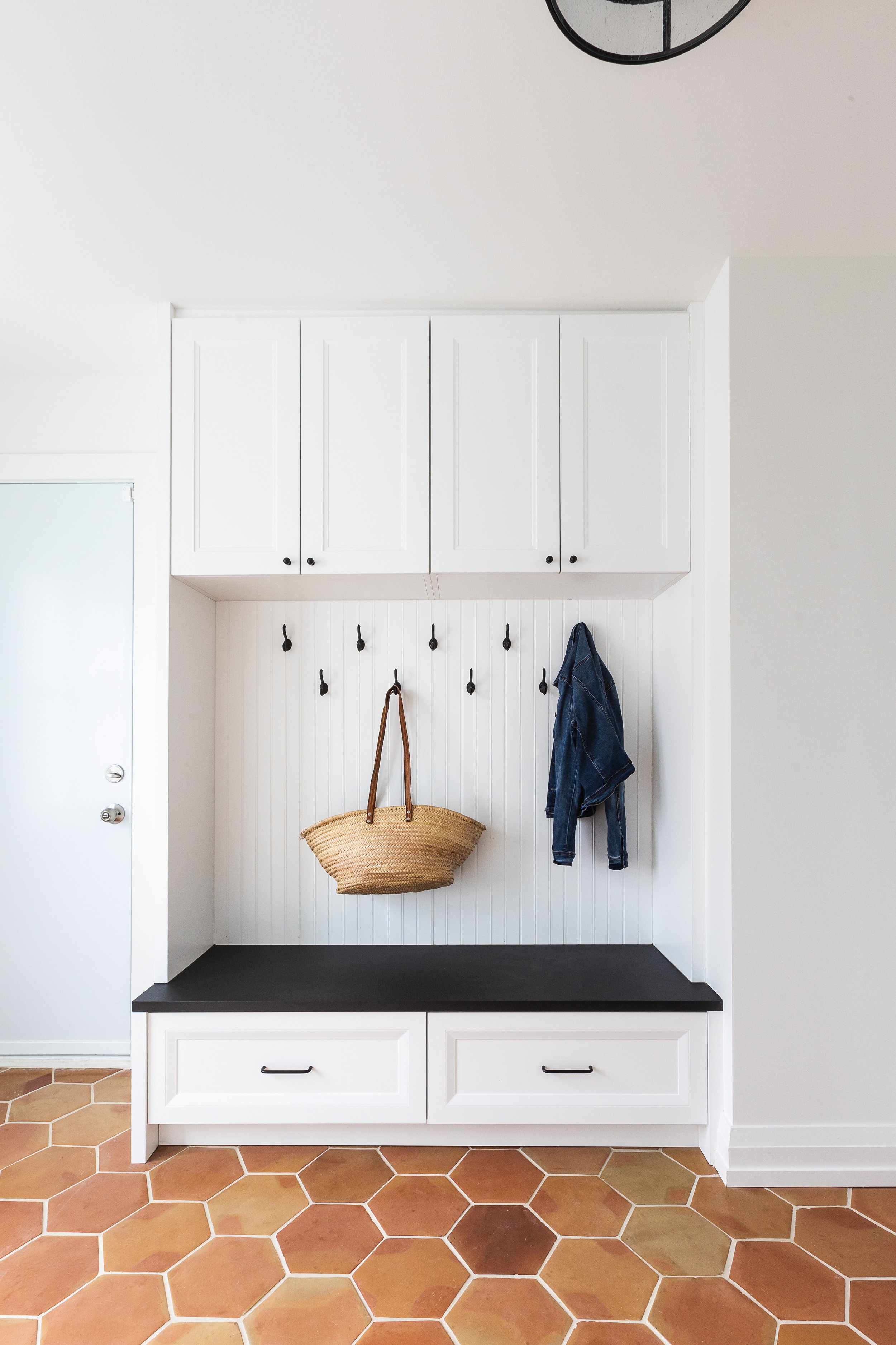 Mudroom with white cabinets, black bench, terracotta hexagonal tiles, coat hooks with denim jacket and straw tote bag.
