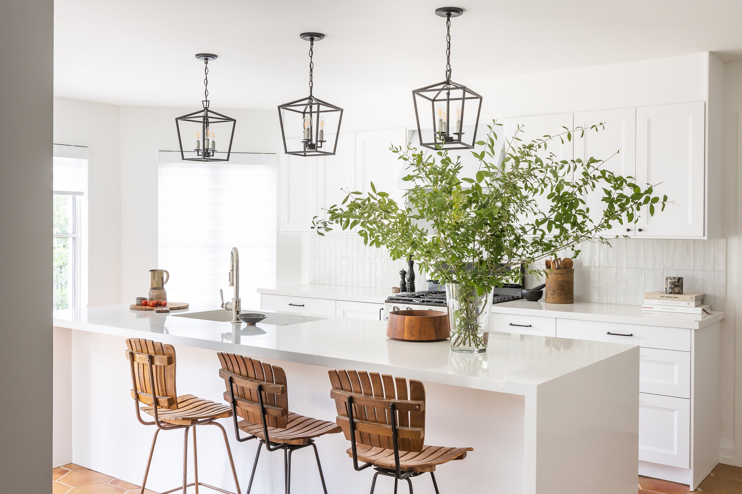 Modern white kitchen with wooden bar stools, pendant lights, and a large plant centerpiece on the island counter.