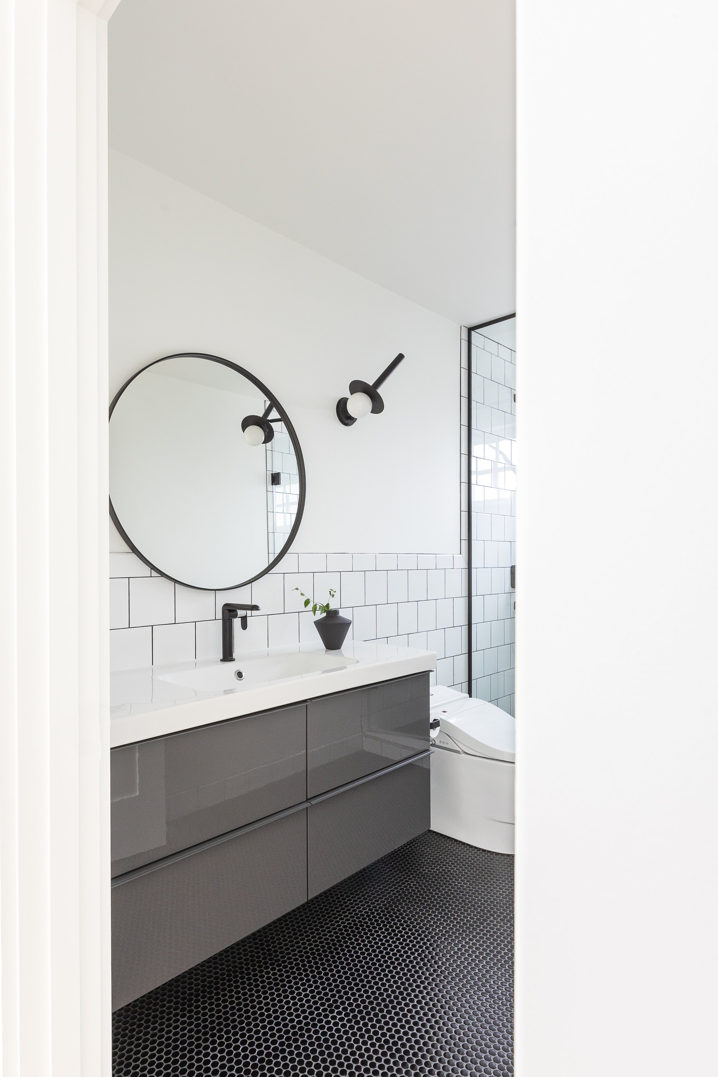 Modern bathroom with circular mirror, black fixtures, white subway tiles, dark vanity, black hexagonal floor tiles, and a small potted plant on the counter.