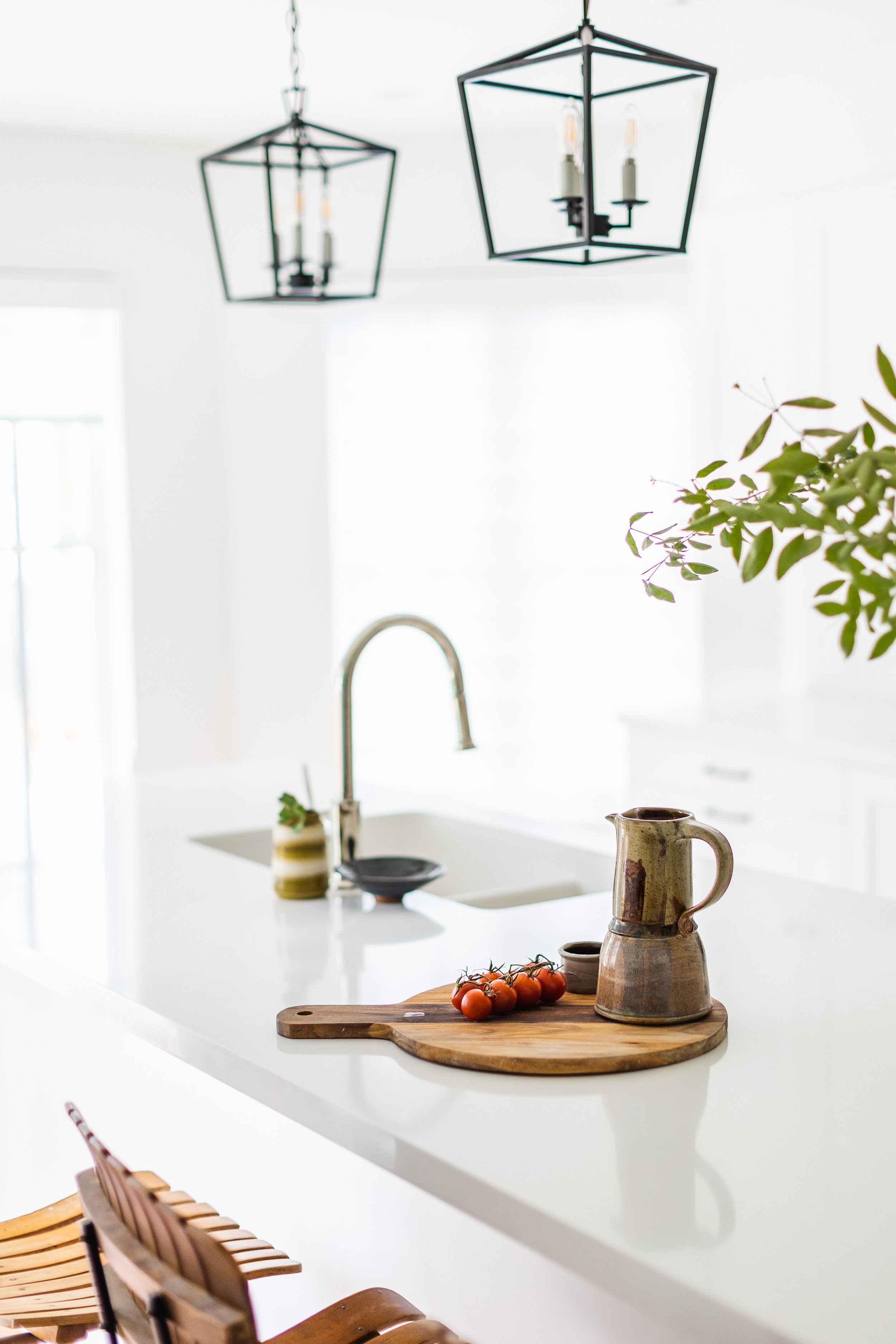 Modern kitchen with white countertop, wooden cutting board with tomatoes, ceramic jug, hanging light fixtures, and indoor plant.