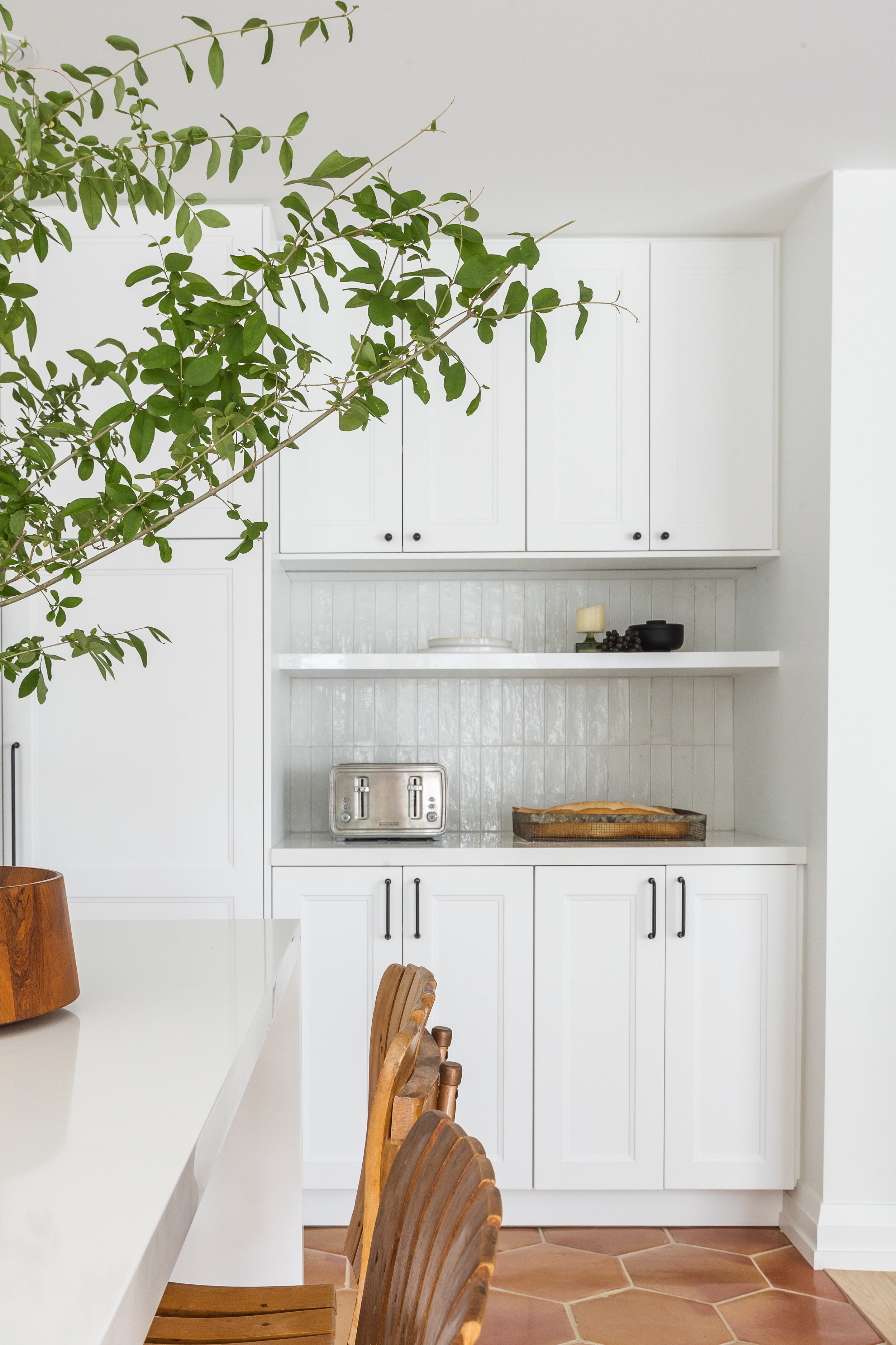 Modern white kitchen with a wooden dining table and chairs, featuring cabinetry, a toaster, a plant arrangement, and decorative shelves.
