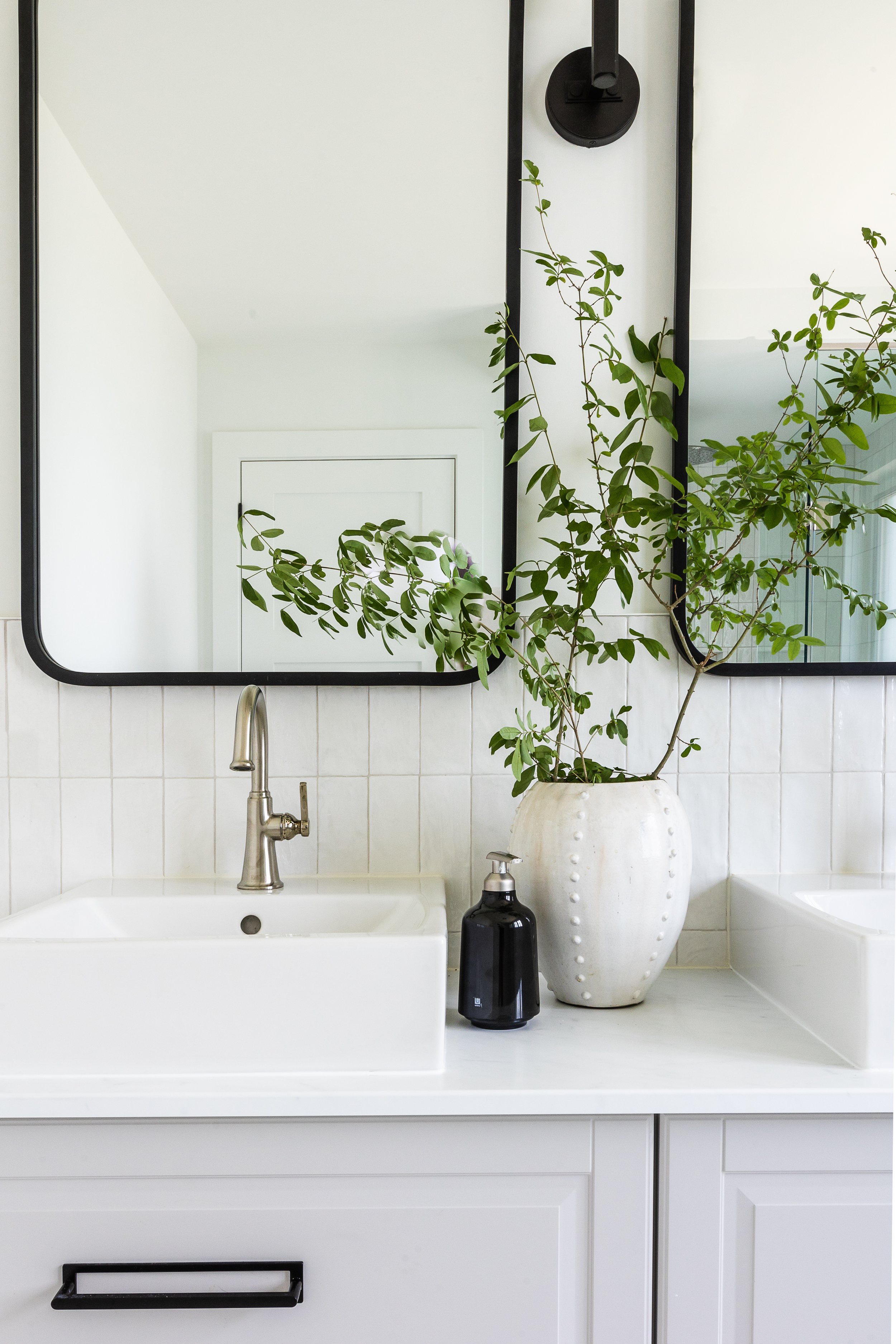 Modern bathroom vanity with white rectangular sink, silver faucet, large rectangular mirror, white vase with green foliage, and black soap dispenser.