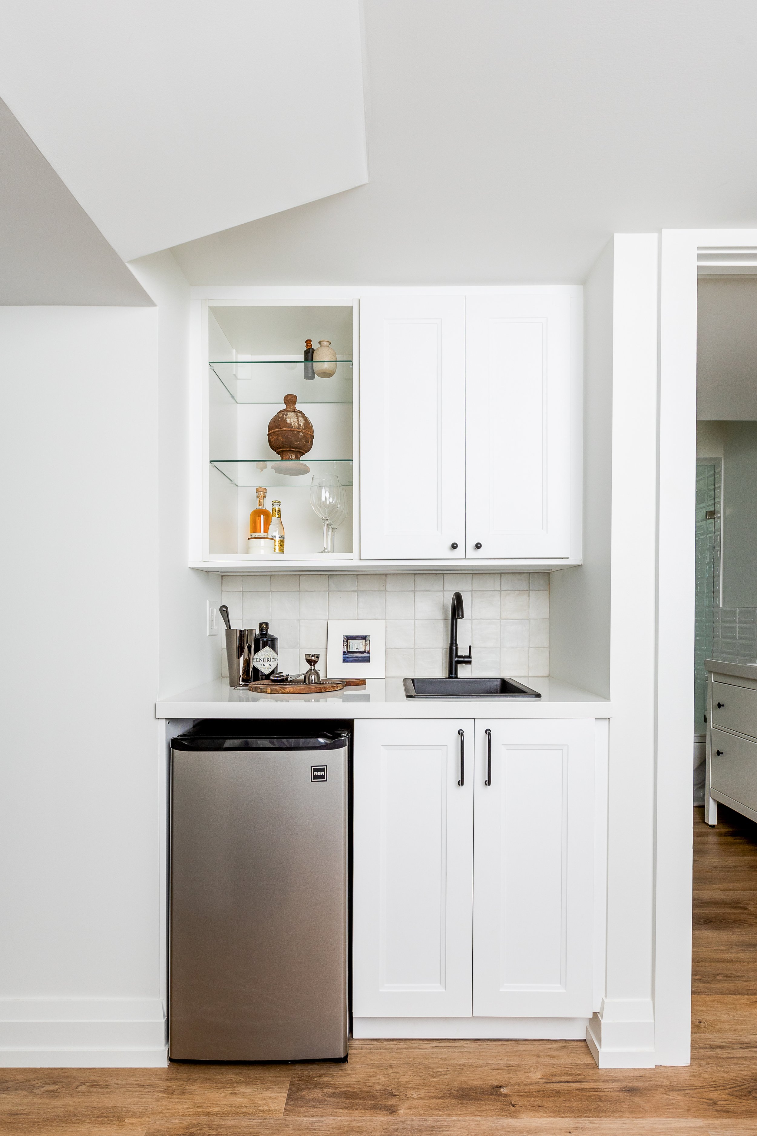 Compact home bar area with white cabinets, small sink, mini fridge, and decorative items on shelves.