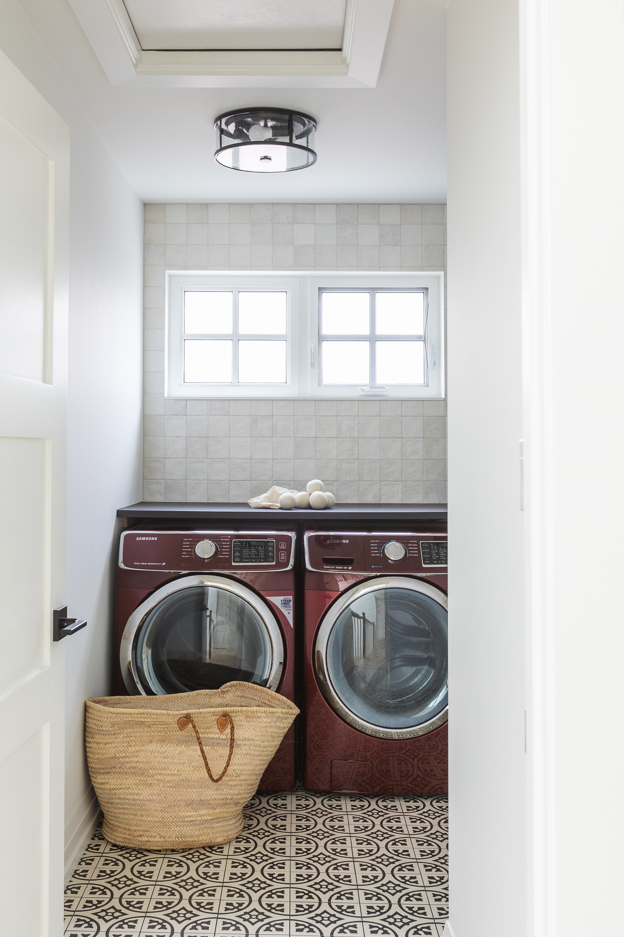Modern laundry room with red washer and dryer, woven basket, tiled floor, and window with natural light.