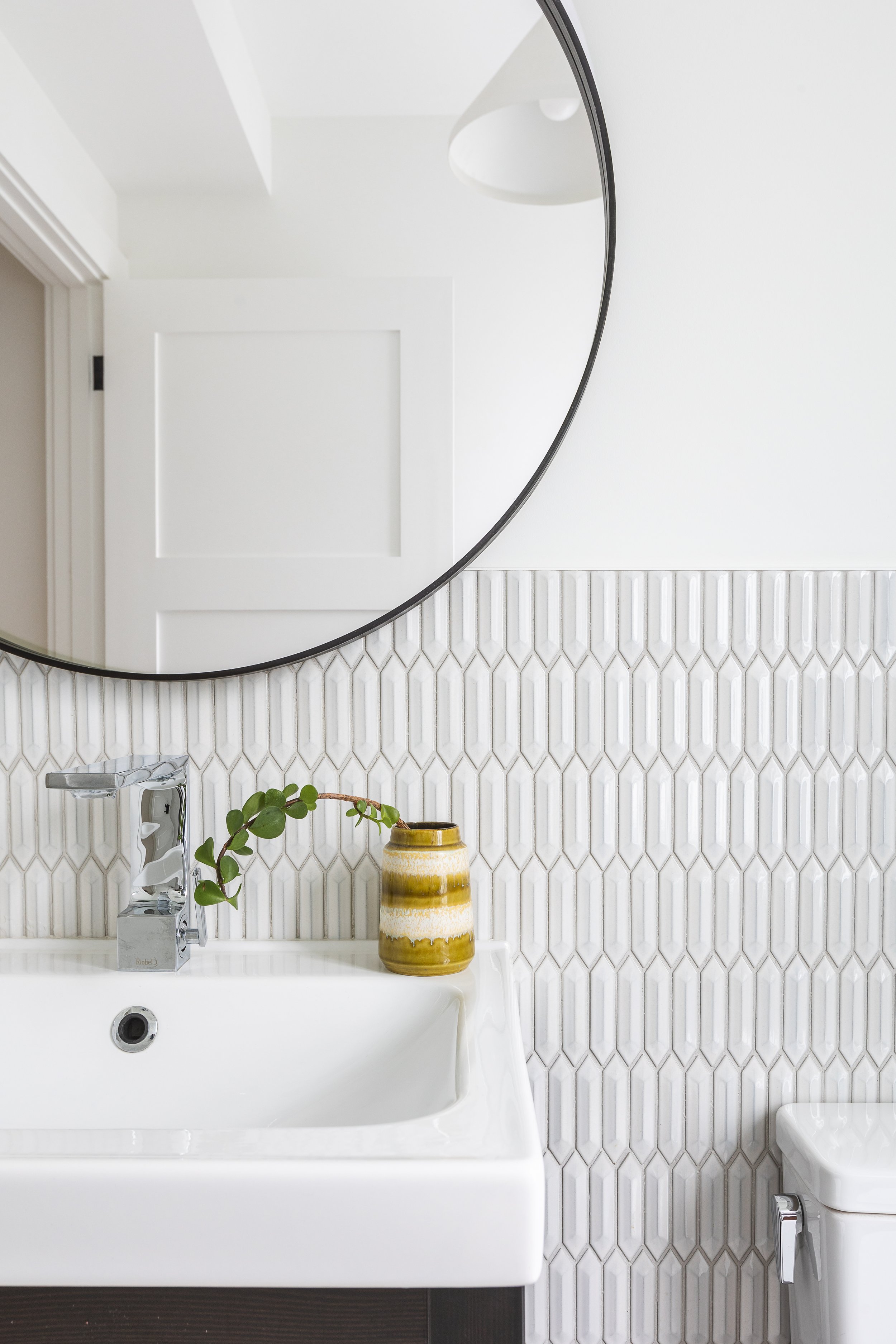 Modern bathroom with round mirror above white sink, geometric tile backsplash, chrome faucet, and decorative plant in ceramic pot.