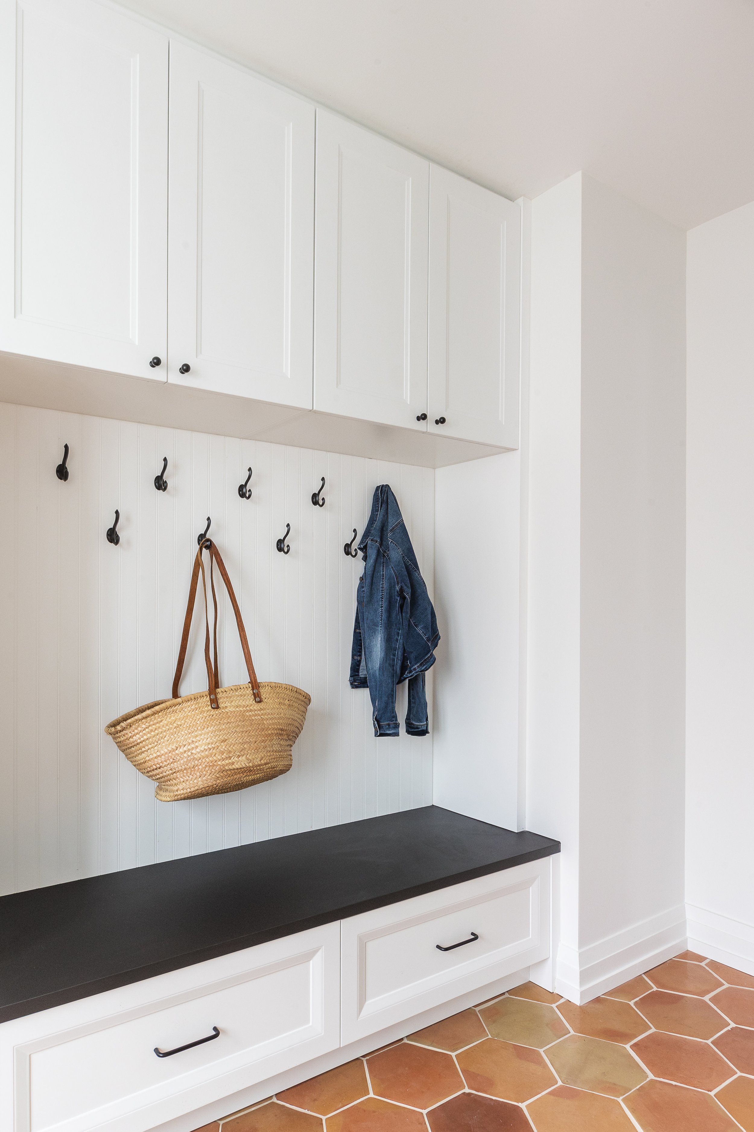 White entryway with hooks, a jean jacket, and woven basket; black bench and cabinets above.