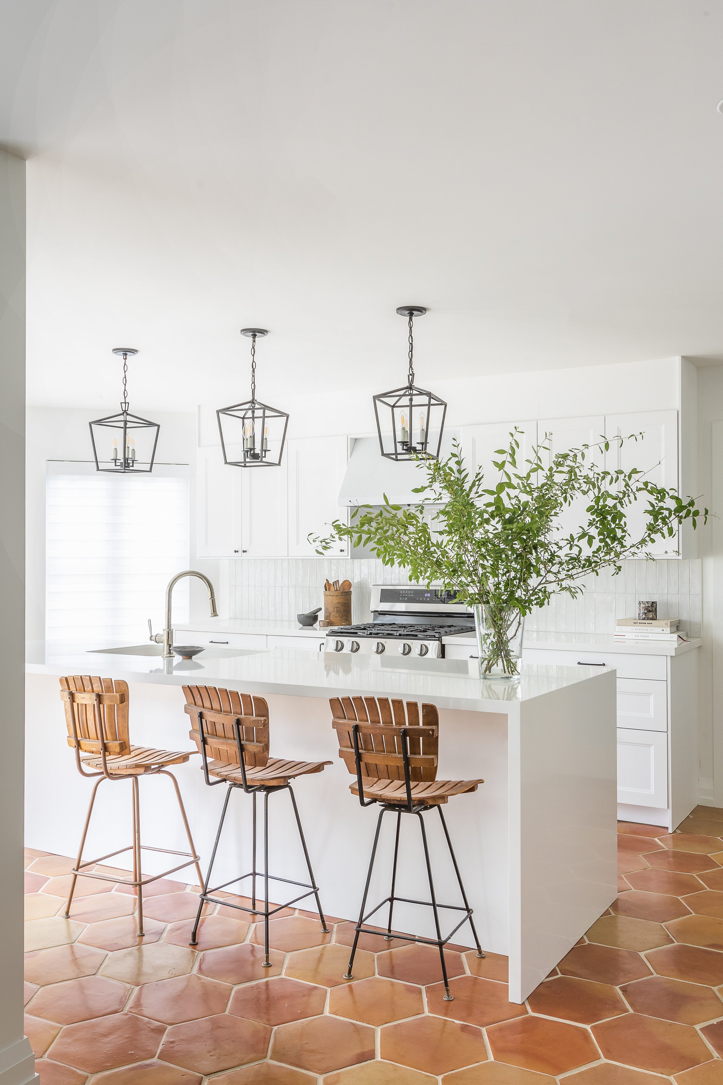 Modern kitchen with white cabinets, a large island, three wooden bar stools, pendant lights, and a vase with greenery on the countertop. Hexagonal tile flooring.