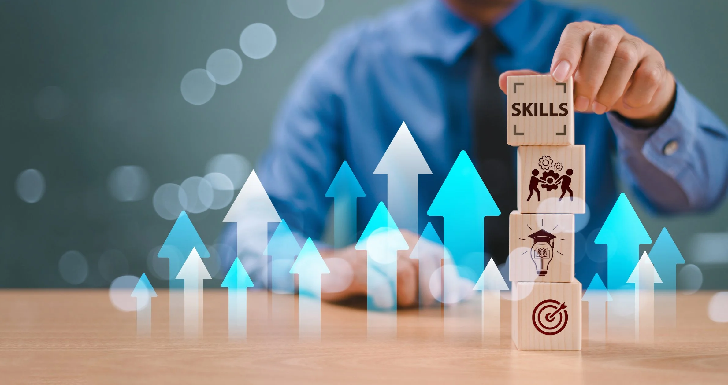 A person in a blue shirt stacking wooden blocks labeled "Skills" with icons representing gears, teamwork, education, and a target on top of a table. Blue and white arrows point upward around the blocks, symbolizing growth or improvement.