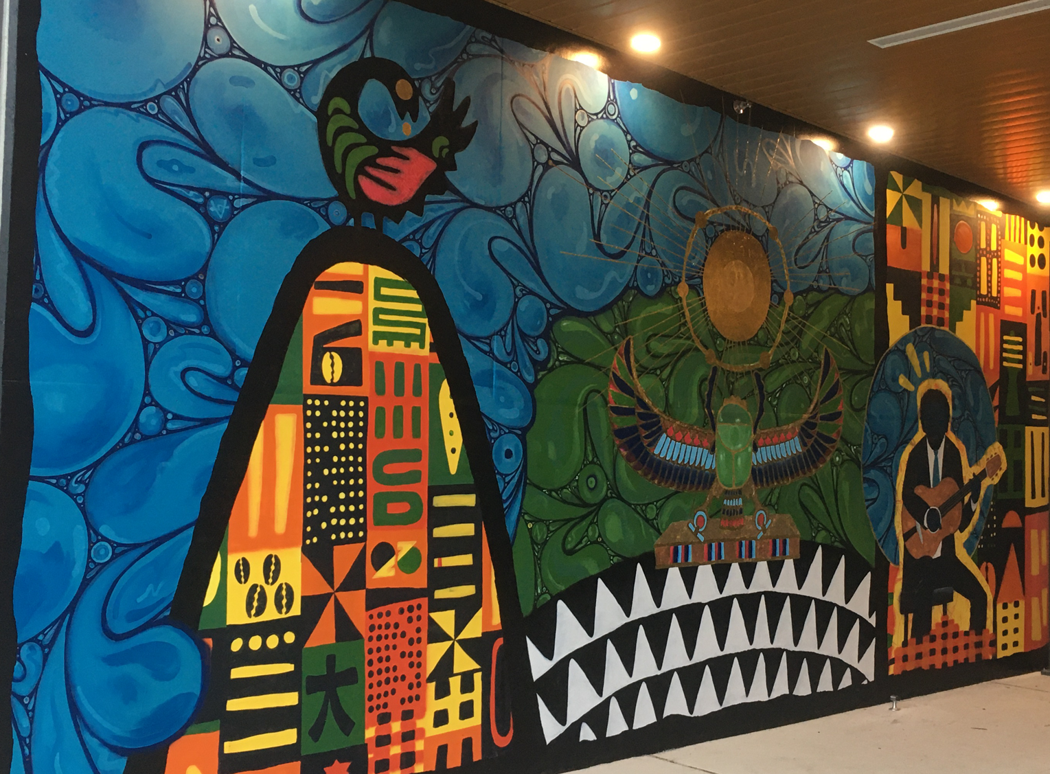 Colorful mural featuring blue abstract shapes, a bird, a rainbow-colored building, a large golden circle, a detailed creature with wings, and a silhouette of a person playing guitar.