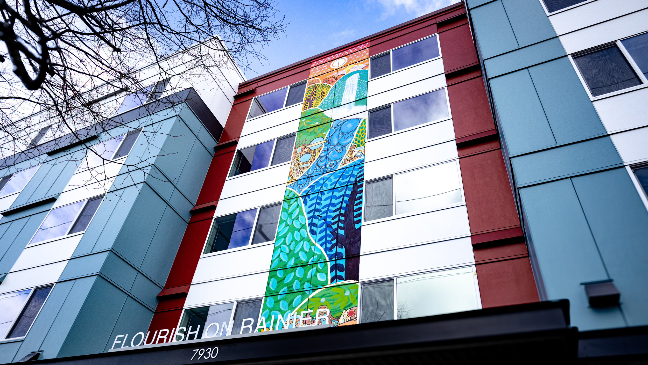 A tall modern building with a colorful mural on its front, featuring abstract landscapes and patterns, with the text 'FLOURISH ON RAINER' and partially visible words at the bottom, and the building's address '7930' below.