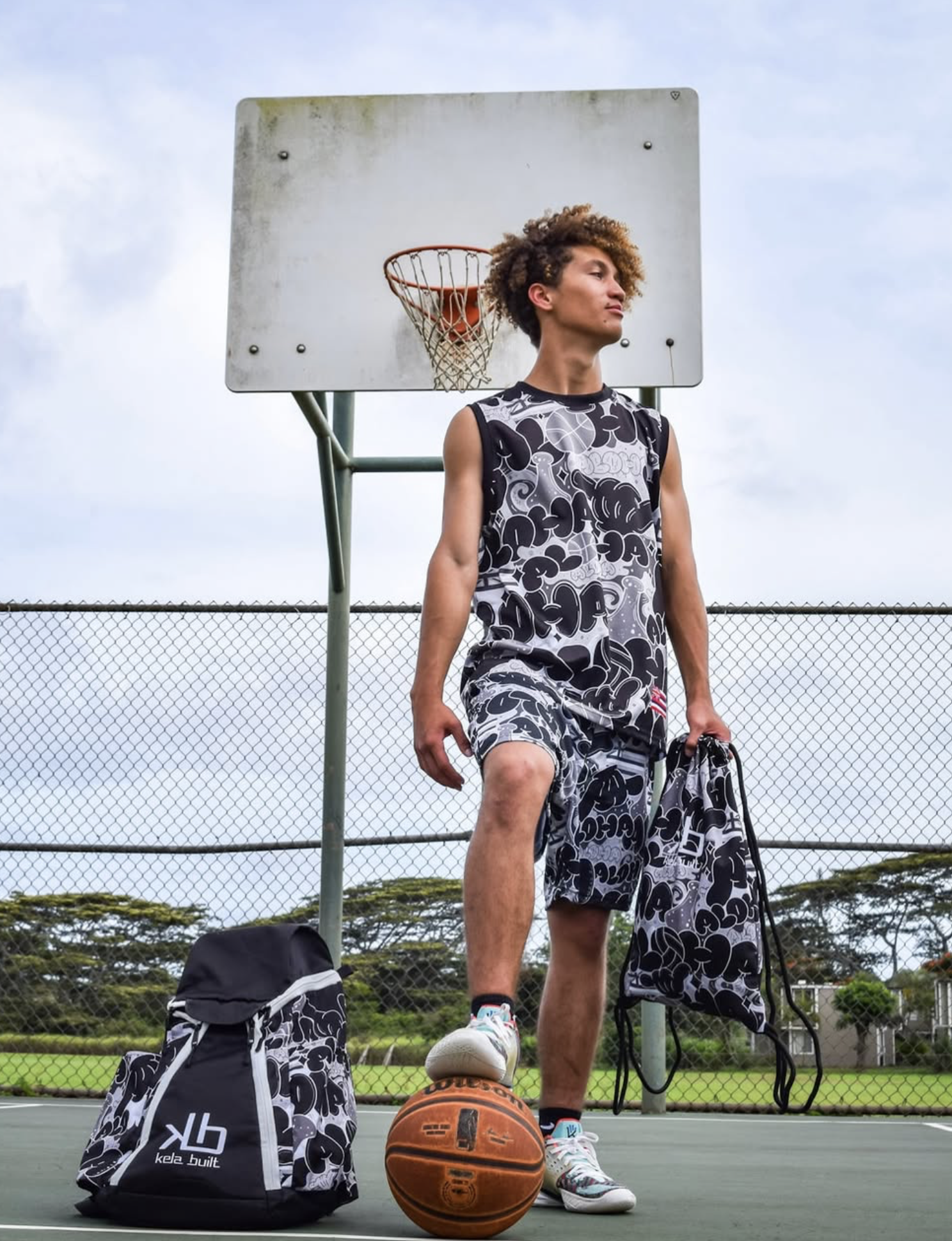 Young man standing on a basketball court with one foot on a basketball, holding a patterned gym bag, wearing matching black and white athletic clothing, near a backpack, in front of a basketball hoop and chain-link fence.