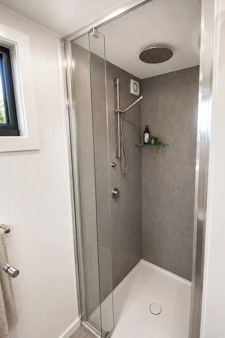 A modern walk-in shower with a glass door, gray tiled walls, a ceiling-mounted rainfall showerhead, a handheld shower with a slide bar, a small glass shelf holding bath products, and a white acrylic floor.