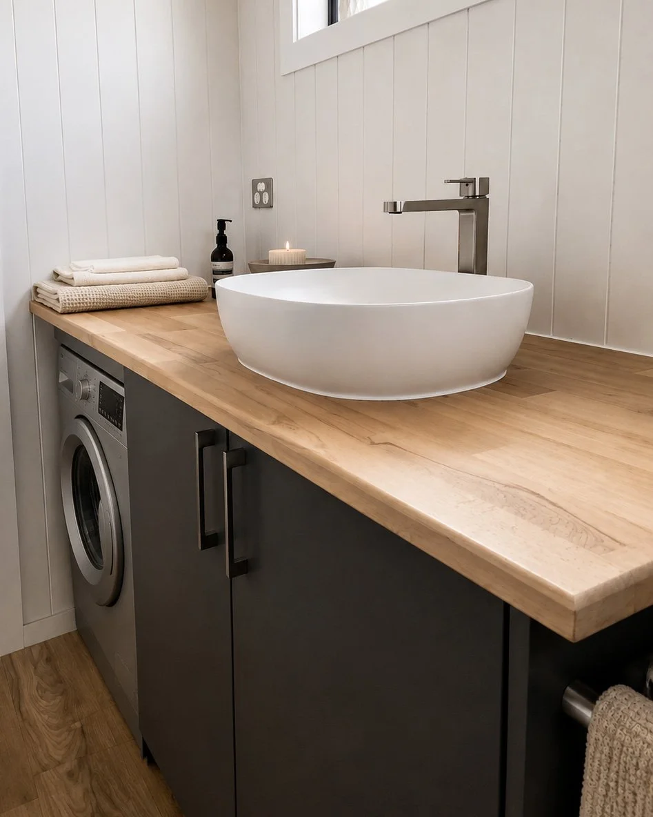 A bathroom vanity with a light wood countertop, a white vessel sink, a tall faucet, a soap dispenser, a candle, and neatly folded towels. There is a washing machine embedded under the counter, and the walls are painted white with wood paneling.