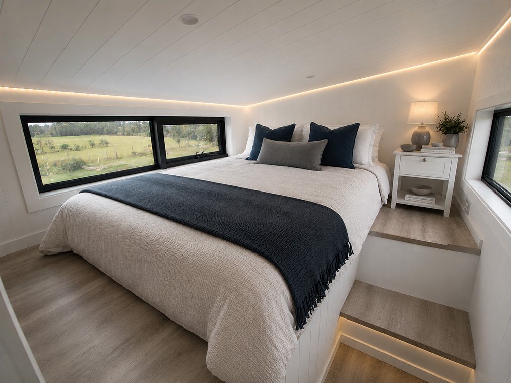 Cozy bedroom with a large bed, dark pillows, a white nightstand with a lamp, and large windows showing a green landscape outside.