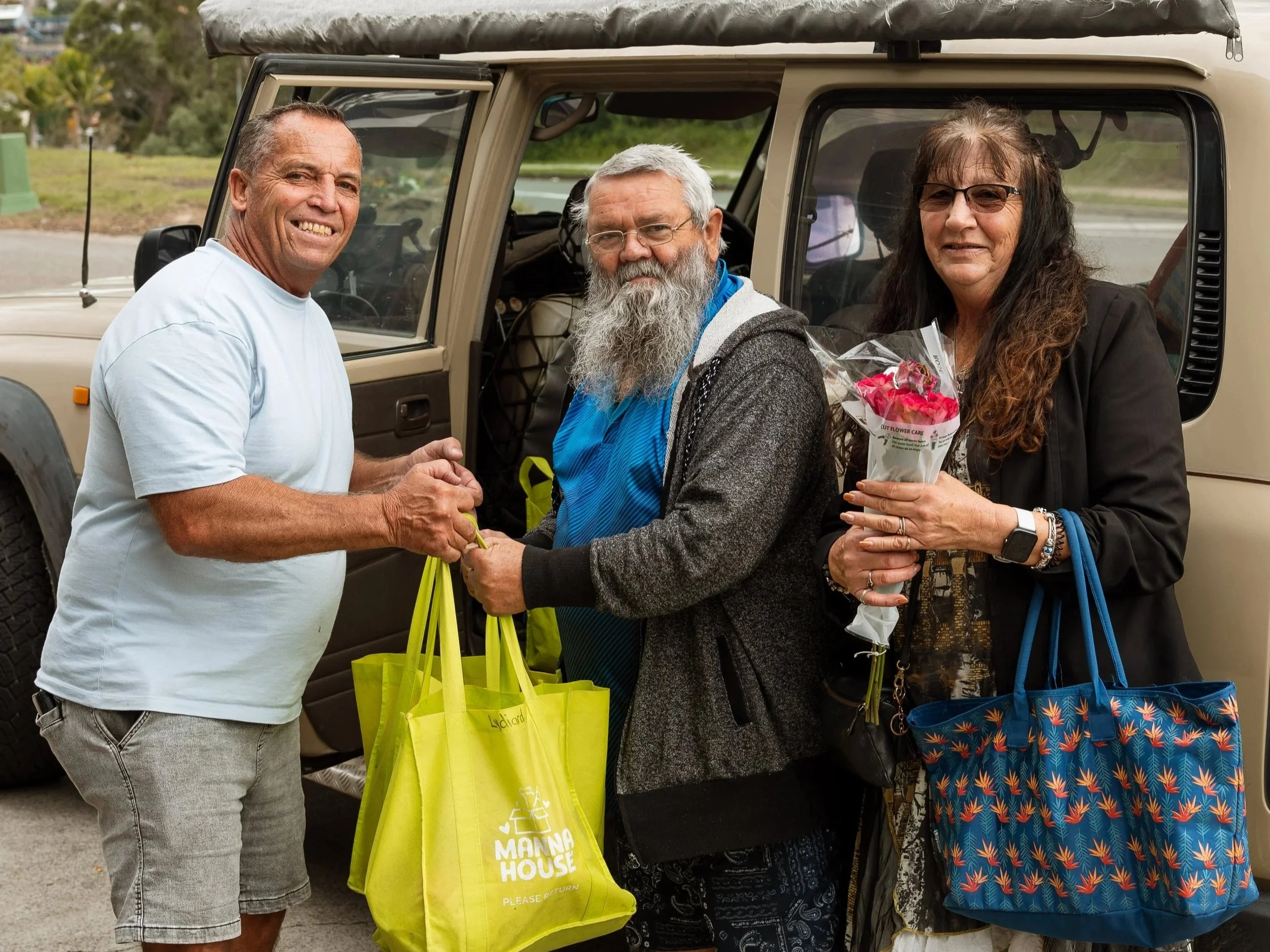 A man and a woman shaking hands in front of a beige off-road vehicle. The woman is holding a bouquet of pink flowers, and both are smiling. The woman has glasses, long dark hair, and is wearing a black jacket. The man has short dark hair and is wearing a white t-shirt. The woman has yellow and blue bags hanging from her shoulder.