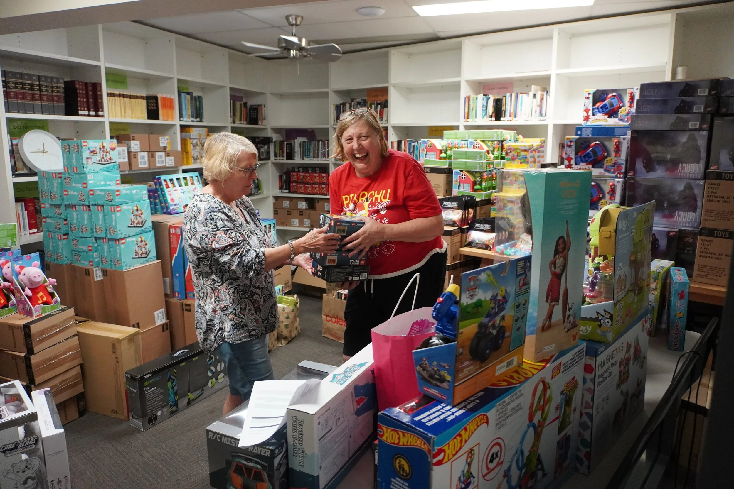 Two women are shopping at a store filled with toys. One woman, wearing glasses and a patterned blouse, is holding and showing a toy to the other woman, who is laughing and wearing a red sweatshirt. The shelves are stocked with various boxes of toys and books.