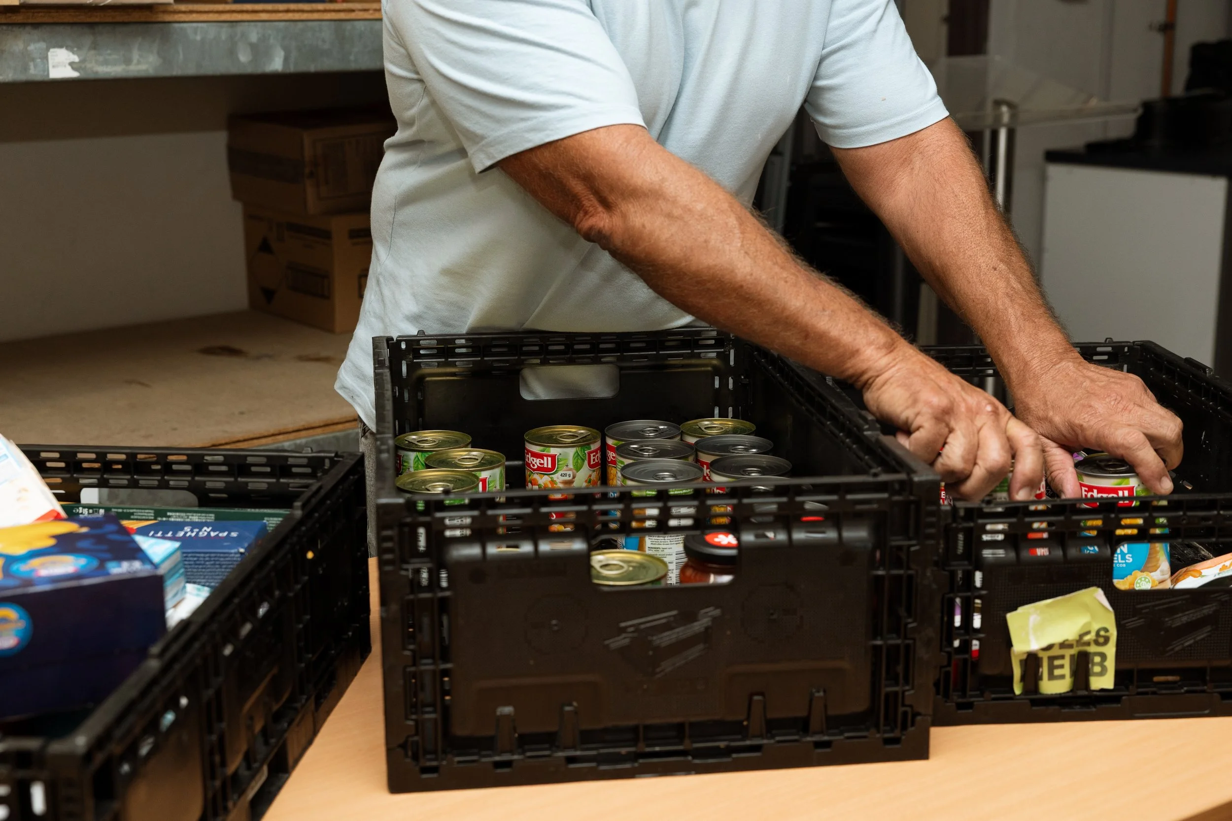 Person wearing a light gray T-shirt packing canned goods into black plastic crates on a table in a storage room.