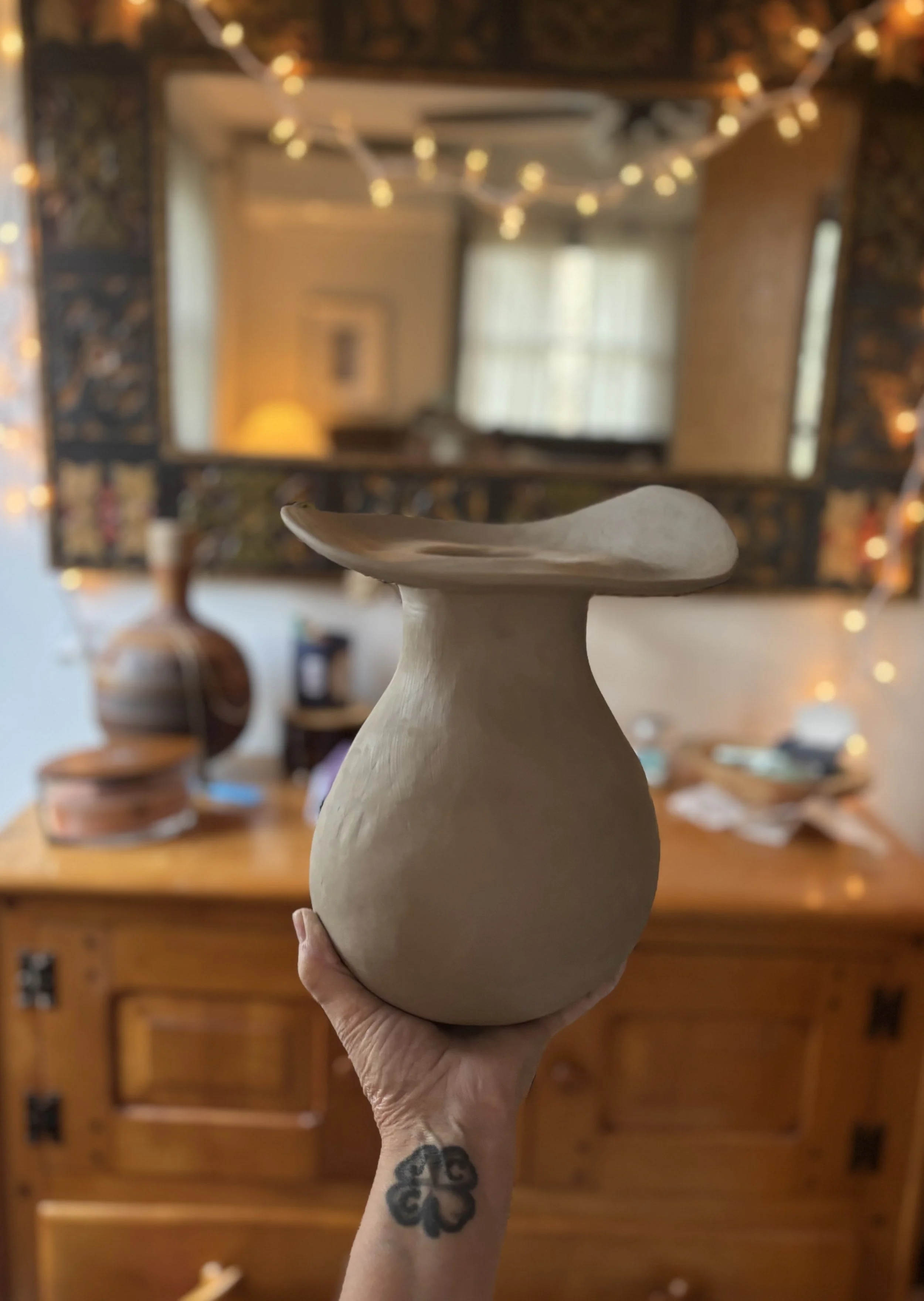 A person holding a neutral-colored ceramic vase with a wide rim in a cozy, decorated room with string lights, a mirror, and a wooden dresser.