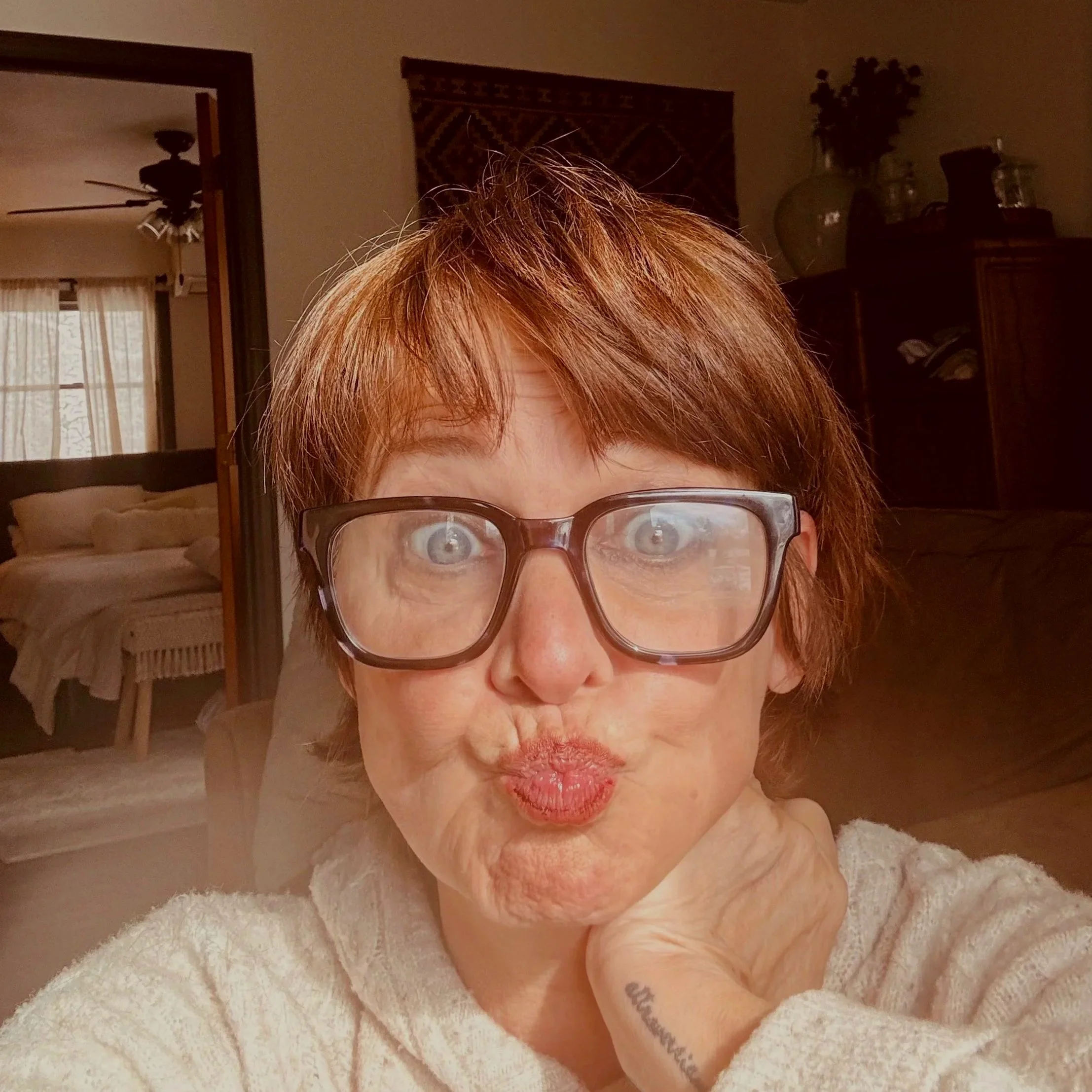 A woman with short reddish hair and glasses making a puckered lips face, sitting indoors with a beige sweater and a tattoo on her wrist.