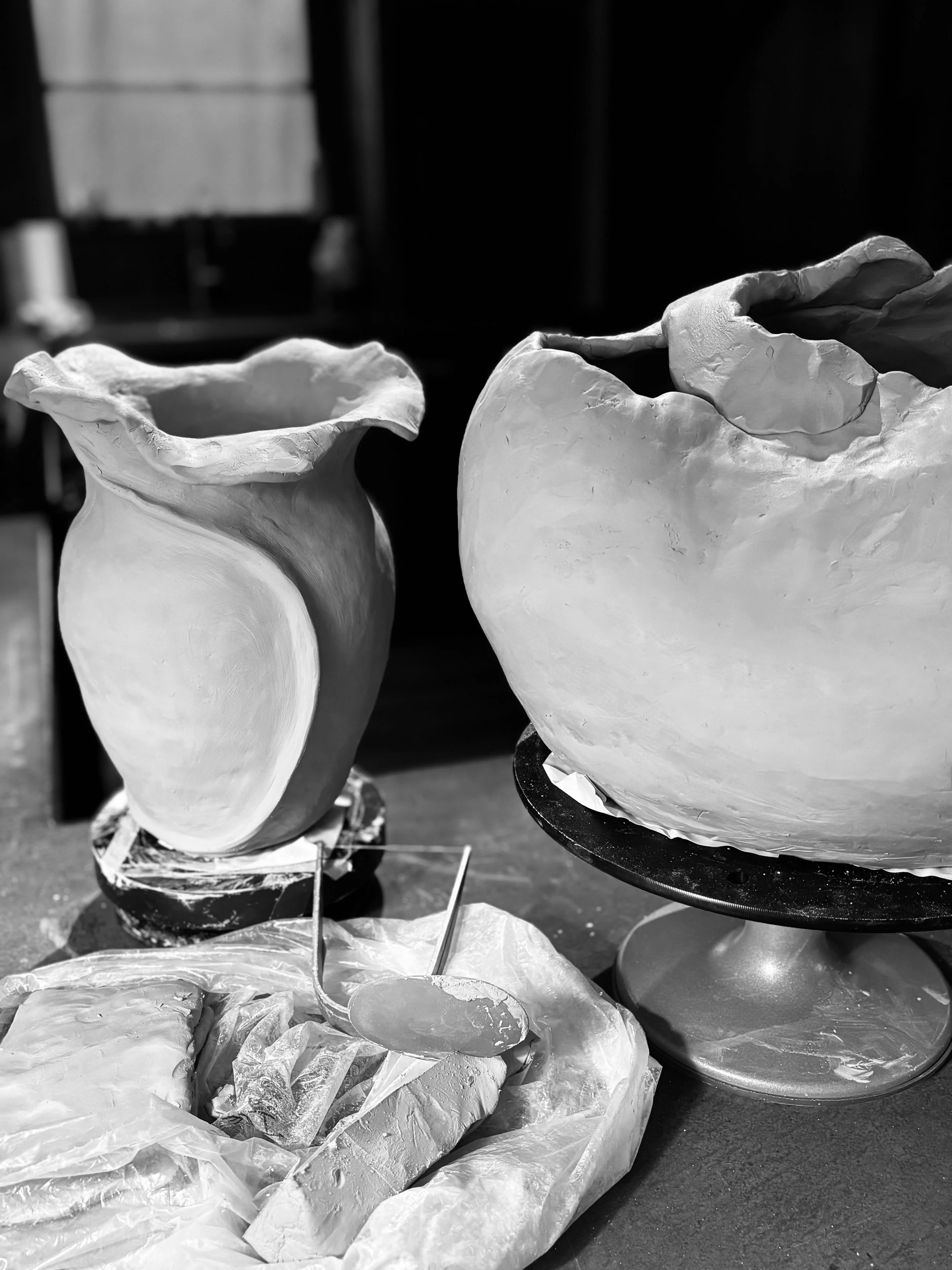 Two unfinished ceramic vases on a table, with tools and clay in the foreground.