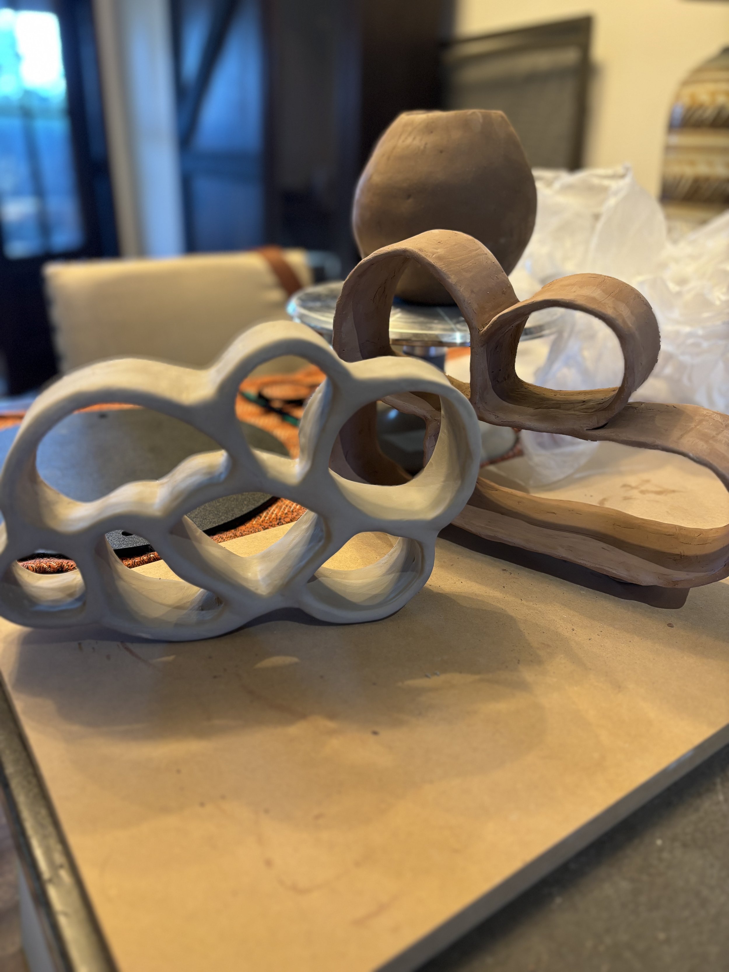 Various handmade pottery and ceramic objects, including a white ceramic piece with honeycomb-like holes, a brown curved handle, an oval-shaped bowl, and a rounded vessel, arranged on a table.