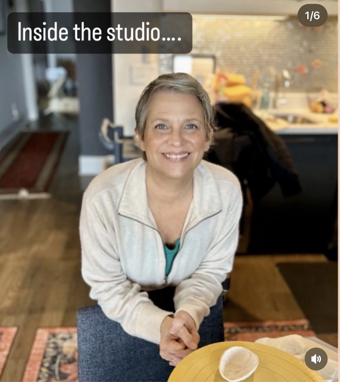 Smiling woman with short gray hair sitting at a table inside a studio or kitchen, with a yellow plate and a bowl in front of her, background shows a kitchen area with various items and a person with long hair bending over.