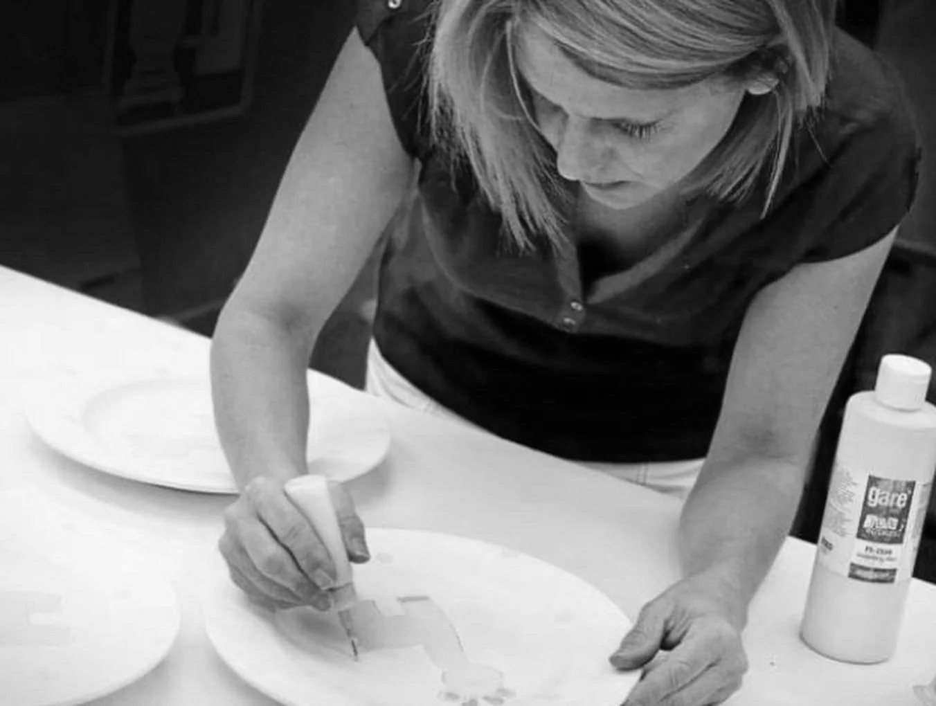A woman is painting on a white ceramic plate with a small brush, with additional plates and a bottle of glaze on the table.