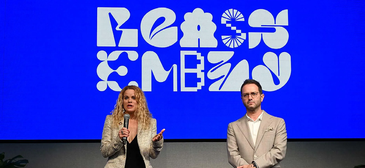 Reassembly brand identity on a blue event screen, with experimental white typography behind two speakers.