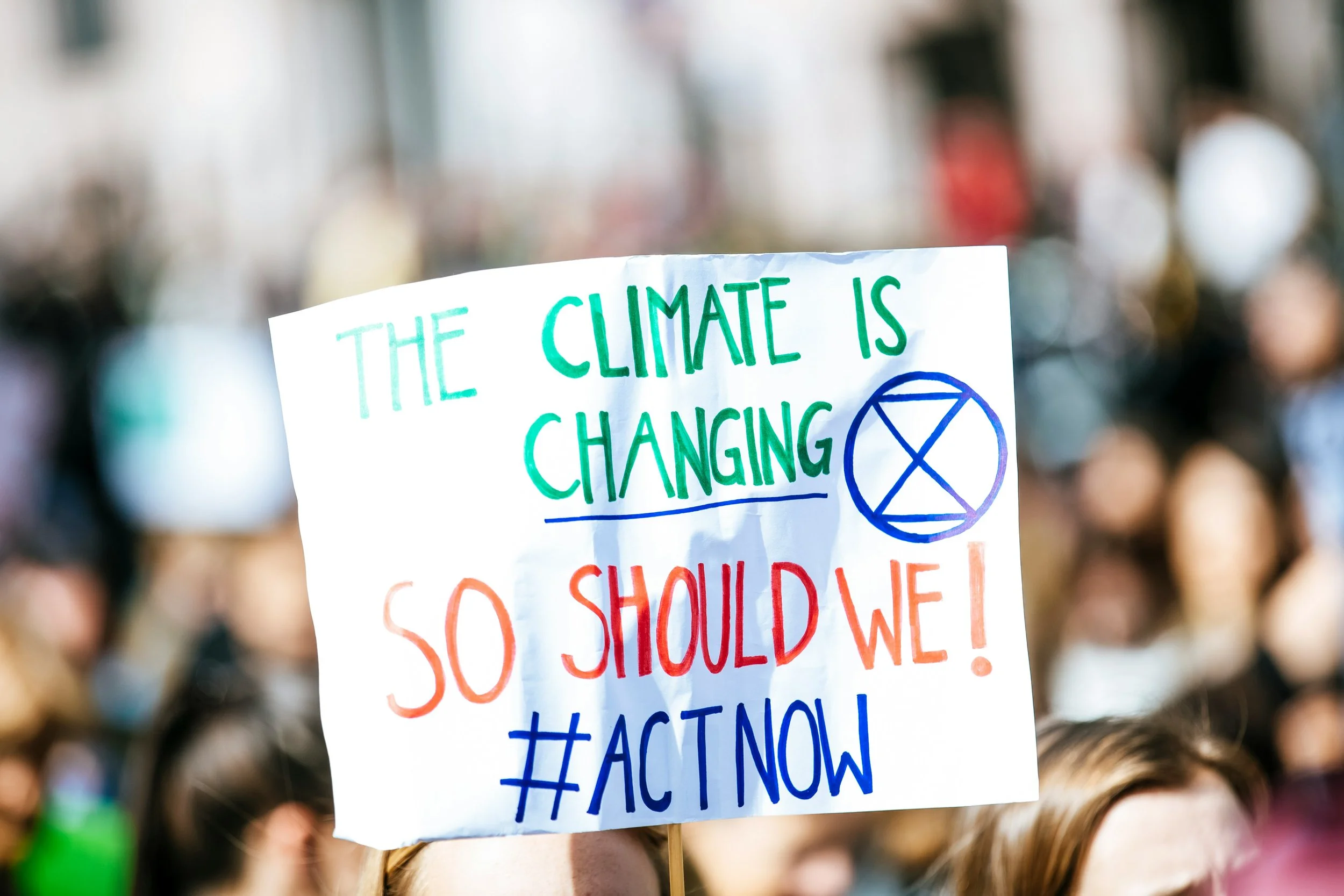 An activist in a crowd holding up a sign that reads "The Climate Is Changing, So Should We! #ACT NOW