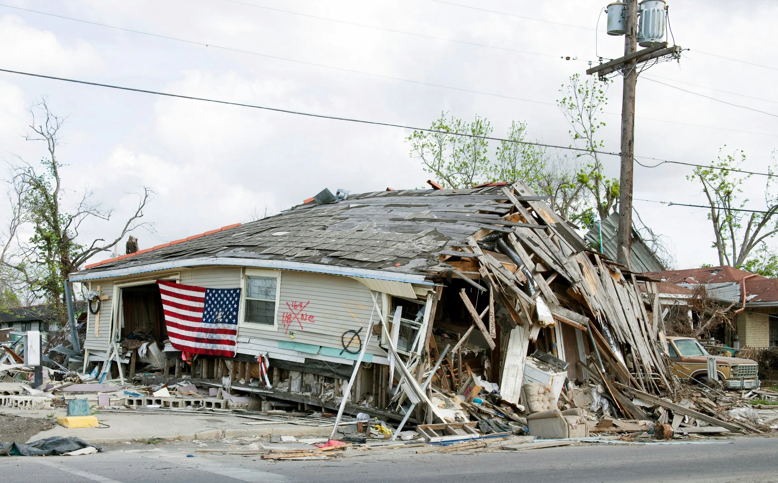 A photo of a home severely damaged by a storm with an American flag hanging on the side of it.