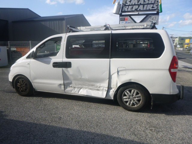 Damaged white van parked in front of a smash repair shop, with visible dents on the side.