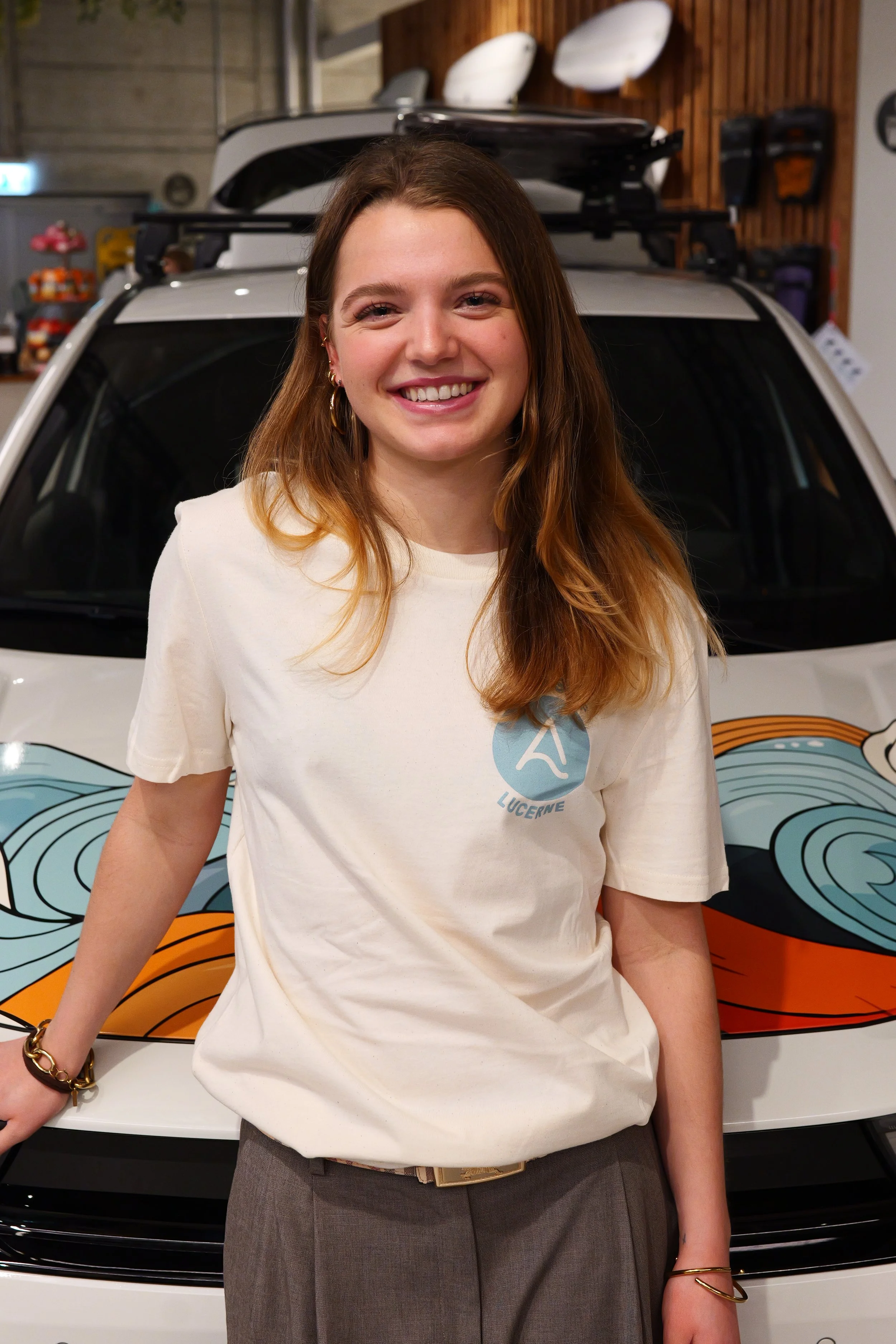 Ein lachendes Mädchen mit lang blondem Haar hält ein Surfbrett mit der Aufschrift "SURFARI" vor einer großen Pflanze in einem Indoor-Bereich.