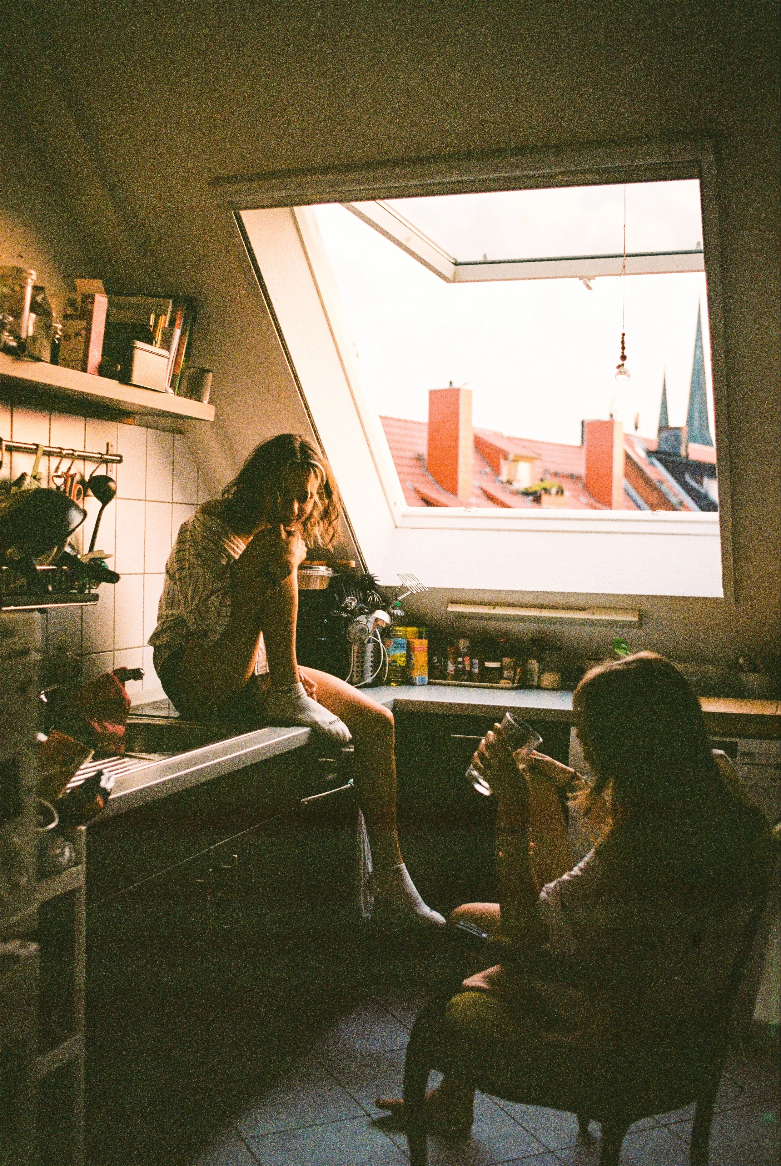 Zwei Mädchen sitzen in einer Dachgeschossküche, eine auf der Küchenspüle und die andere auf einem Stuhl, mit Blick auf ein großes, geöffnetes Dachfenster, durch das man Dächer und Türme der Stadt sieht.