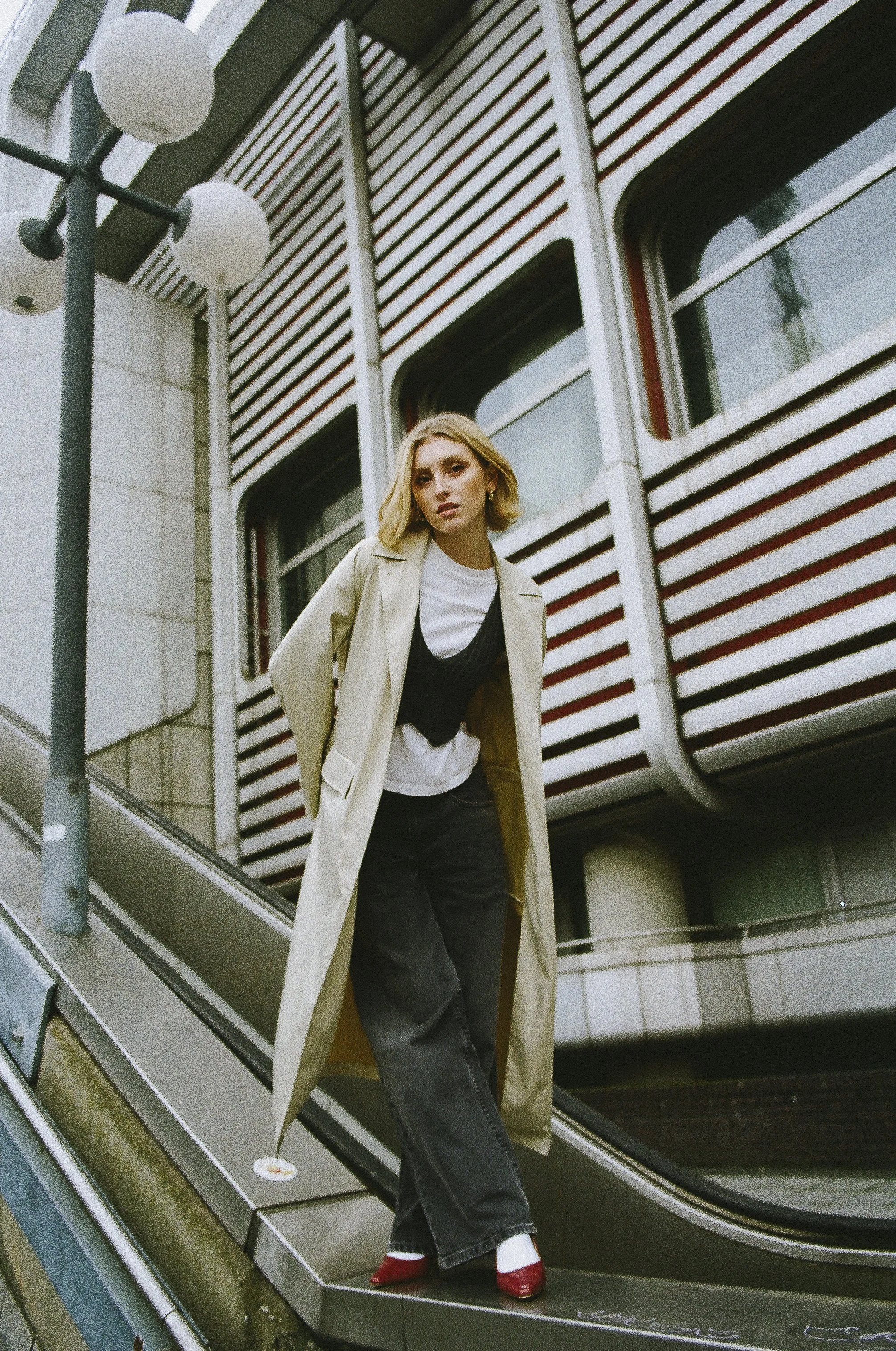 Junge Frau in trennender Jacke, schwarzem Pullover, weißen T-Shirt, schwarzen Jeans und roten Schuhen steht auf einer Rolltreppe vor einem modernen Gebäude mit roten und weißen Balkonen.