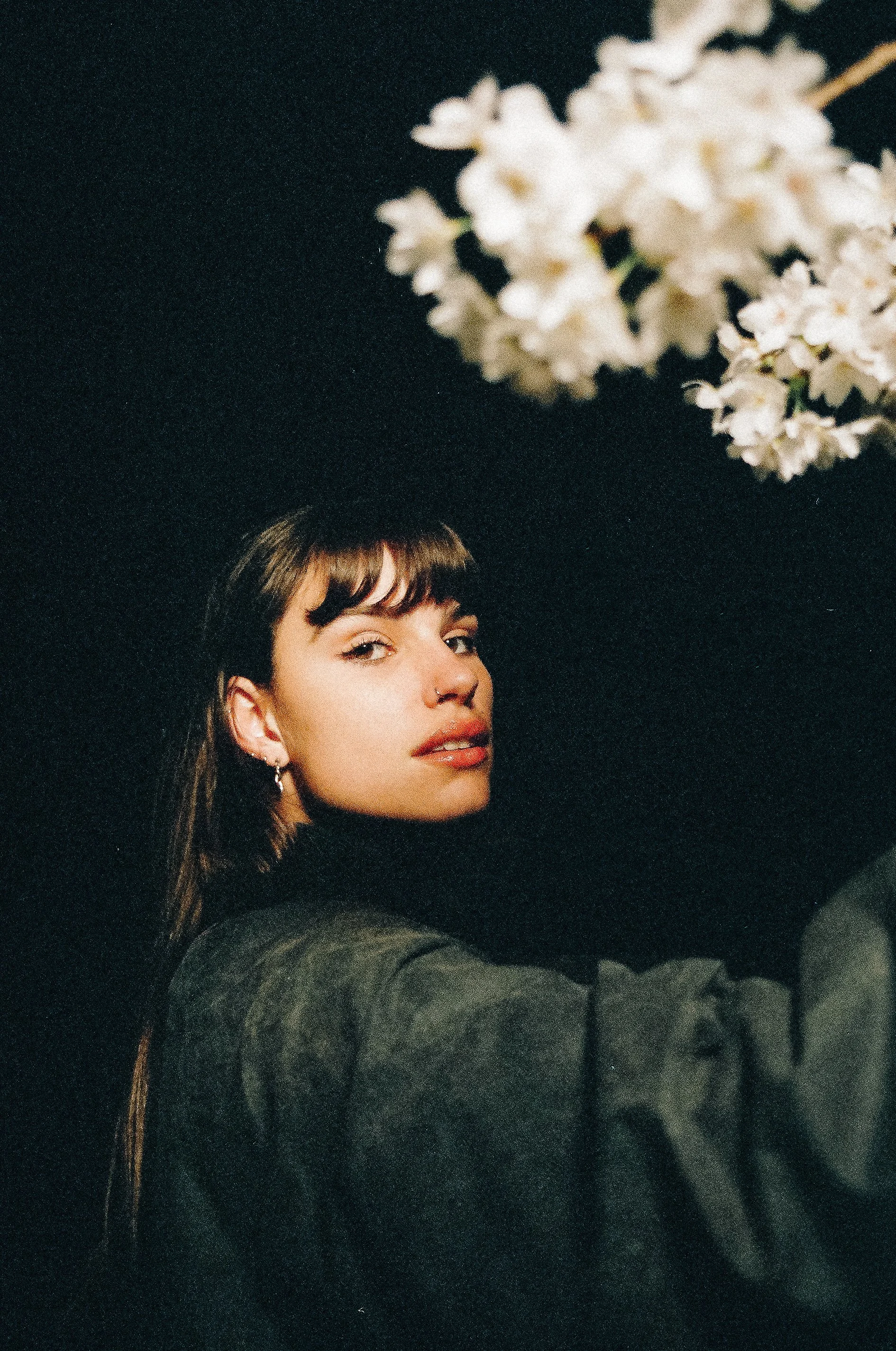 Eine junge Frau mit langen braunen Haaren, grauem Pullover und Ohrringen, posiert vor einem schwarzen Hintergrund, während sie eine weiße Blume hält.
