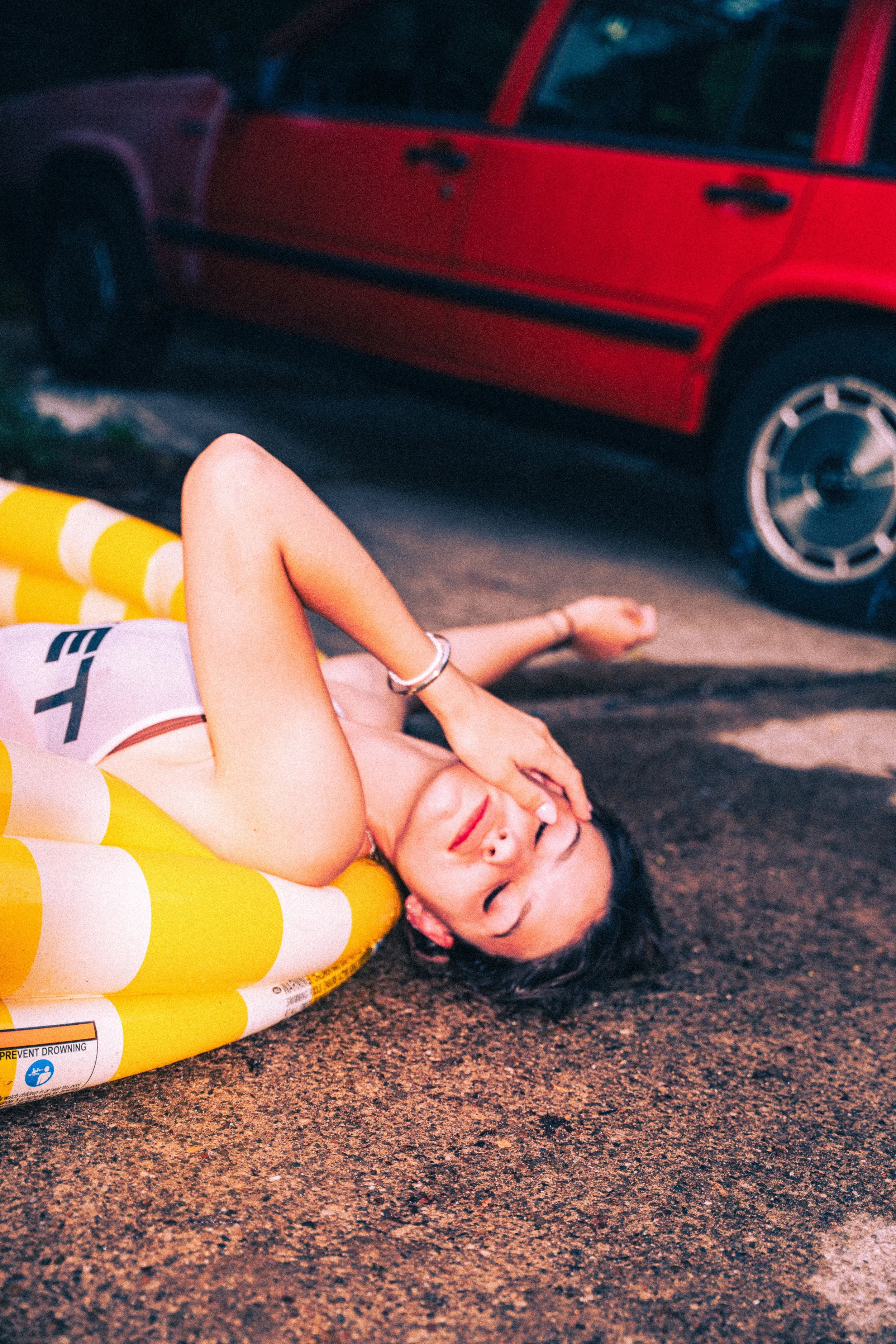 Junge Frau liegt auf dem Boden neben einem aufblasbaren Pool, mit einem roten Auto im Hintergrund, während sie die Augen geschlossen hat.