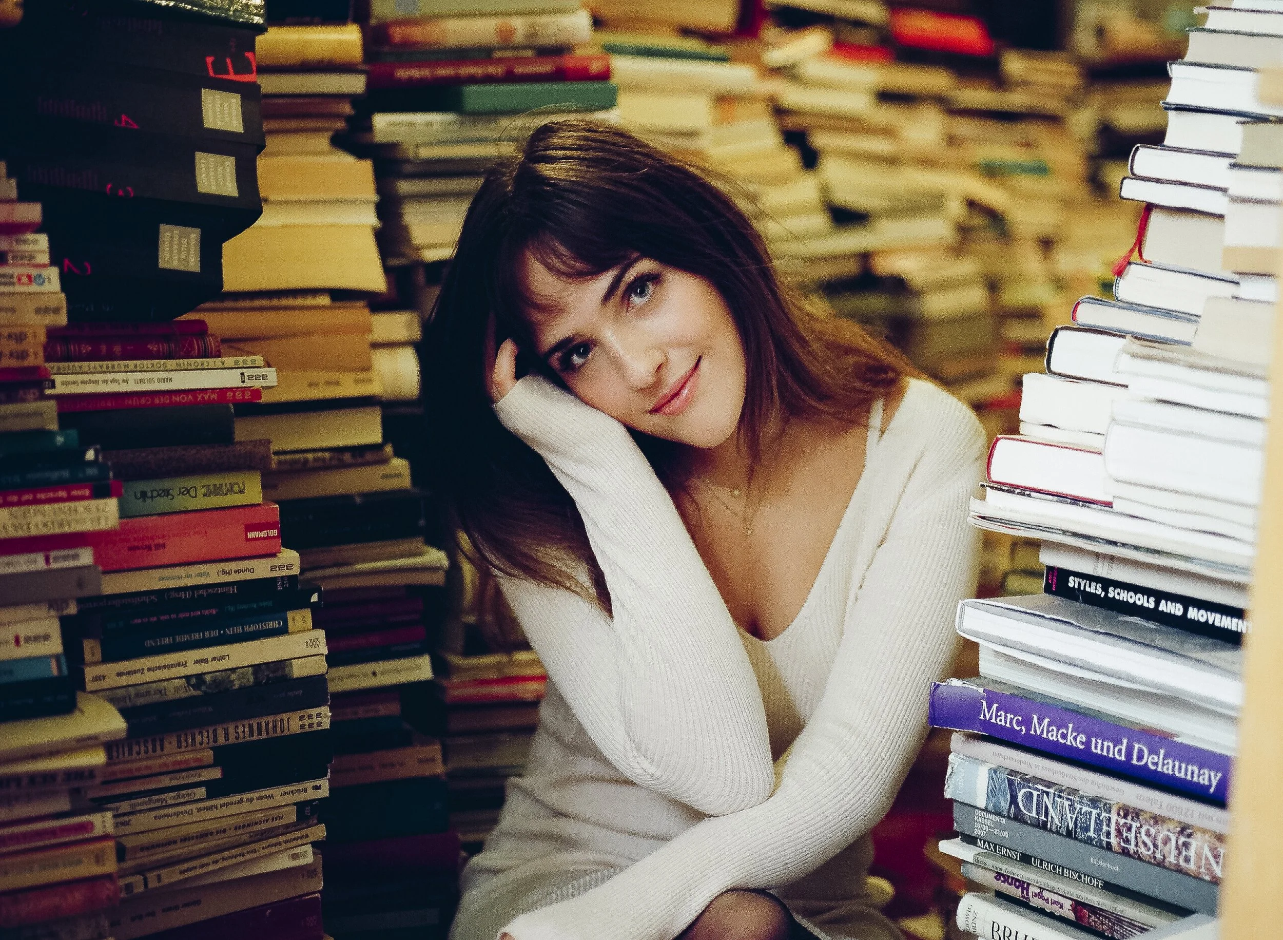 Eine junge Frau sitzt zwischen hohen Bücherstapeln, trägt ein weißes Oberteil und lächelt. Sie hat braune Haare und schaut freundlich in die Kamera.