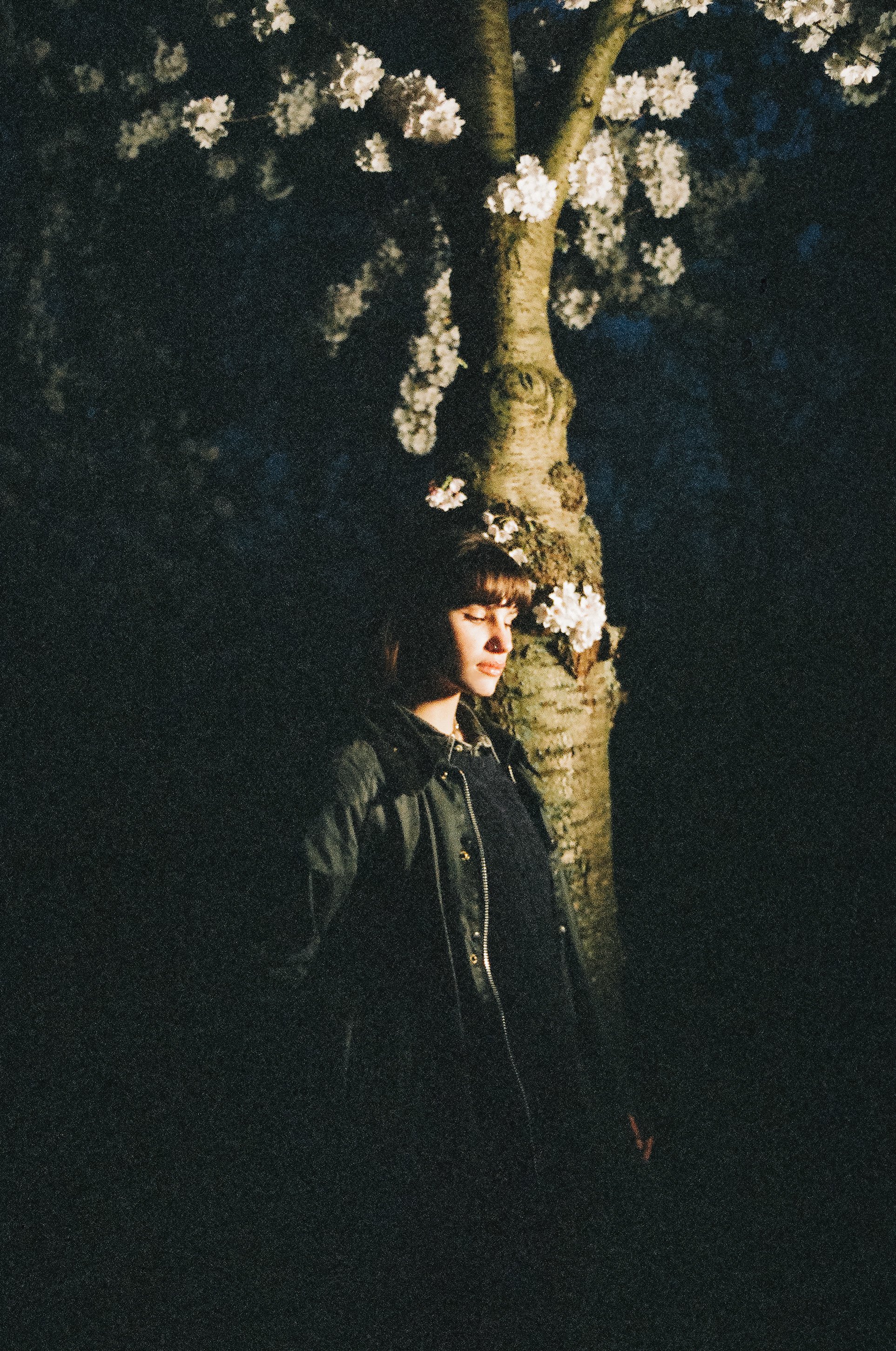 Eine junge Frau mit dunklem Haar steht bei Nacht neben einem blühenden Baum, die Augen geschlossen und vom Licht beleuchtet.
