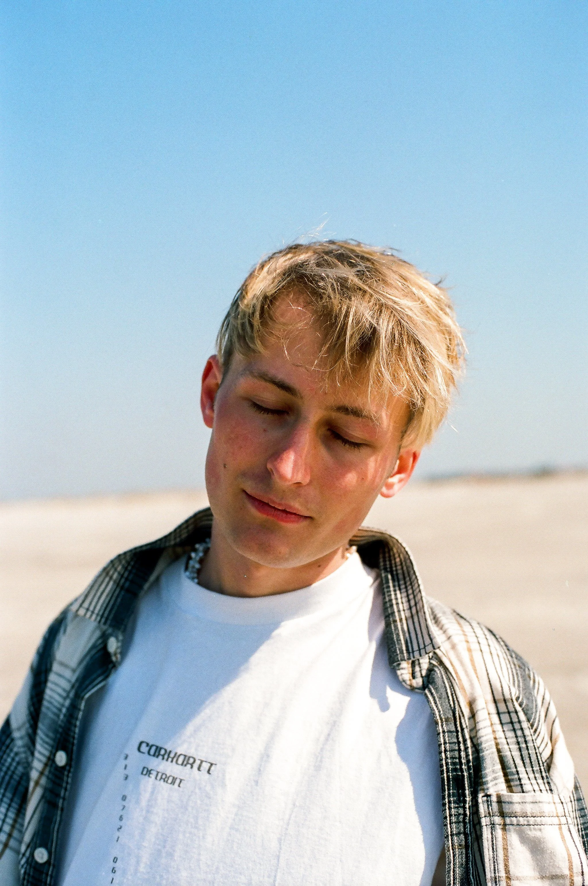 Ein junger Mann mit blondem Haar, der im Sonnenschein mit geschlossenen Augen an einem Strand steht.