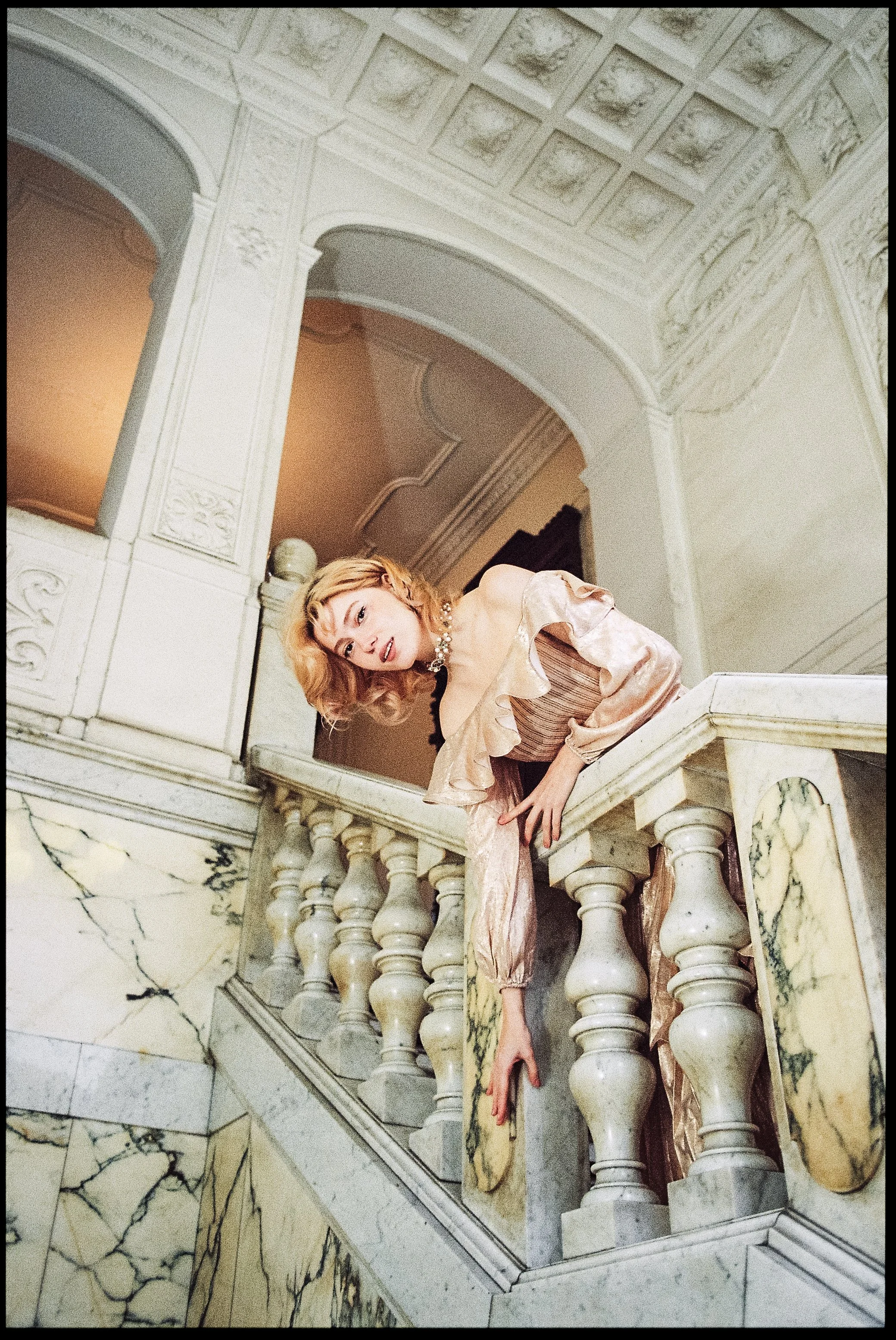 Eine Frau in einem eleganten, seidigen, pastellfarbenen Kleid lehnt an einem gelb- und grün gemarbten Marmorfass auf einer prächtigen Neoklassischen Marmortreppe, umgeben von kunstvoll verzierten weißen Wänden und Decken im Stil des Barock oder Rokok