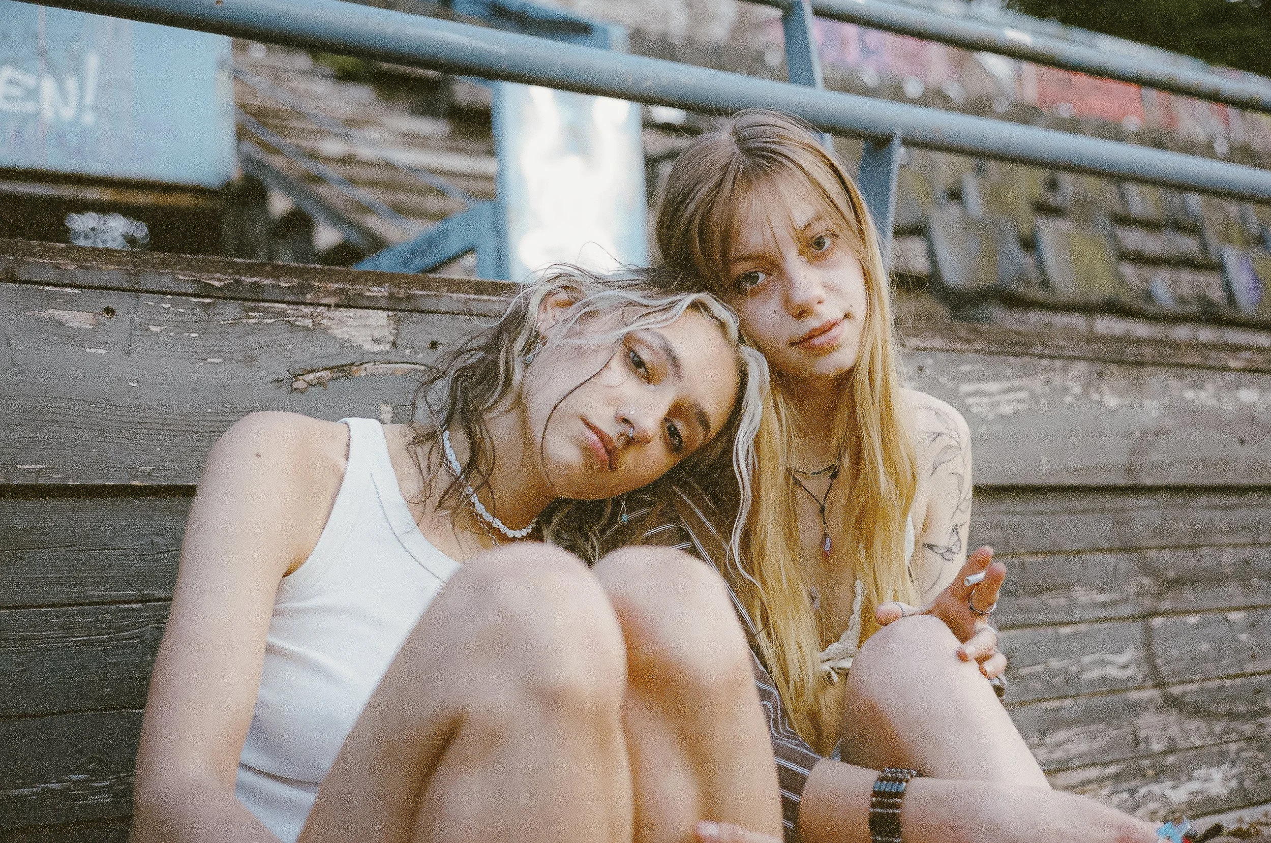 Zwei junge Frauen sitzen auf einer Holzbank im Freien, eine hat lockige, blond-weiß gefärbte Haare, trägt eine weiße Weste und mehrere Halsketten, die andere hat lange, gerade, blondes Haar, trägt ein grünes Top und Schmuck, und sie schauen entspannt