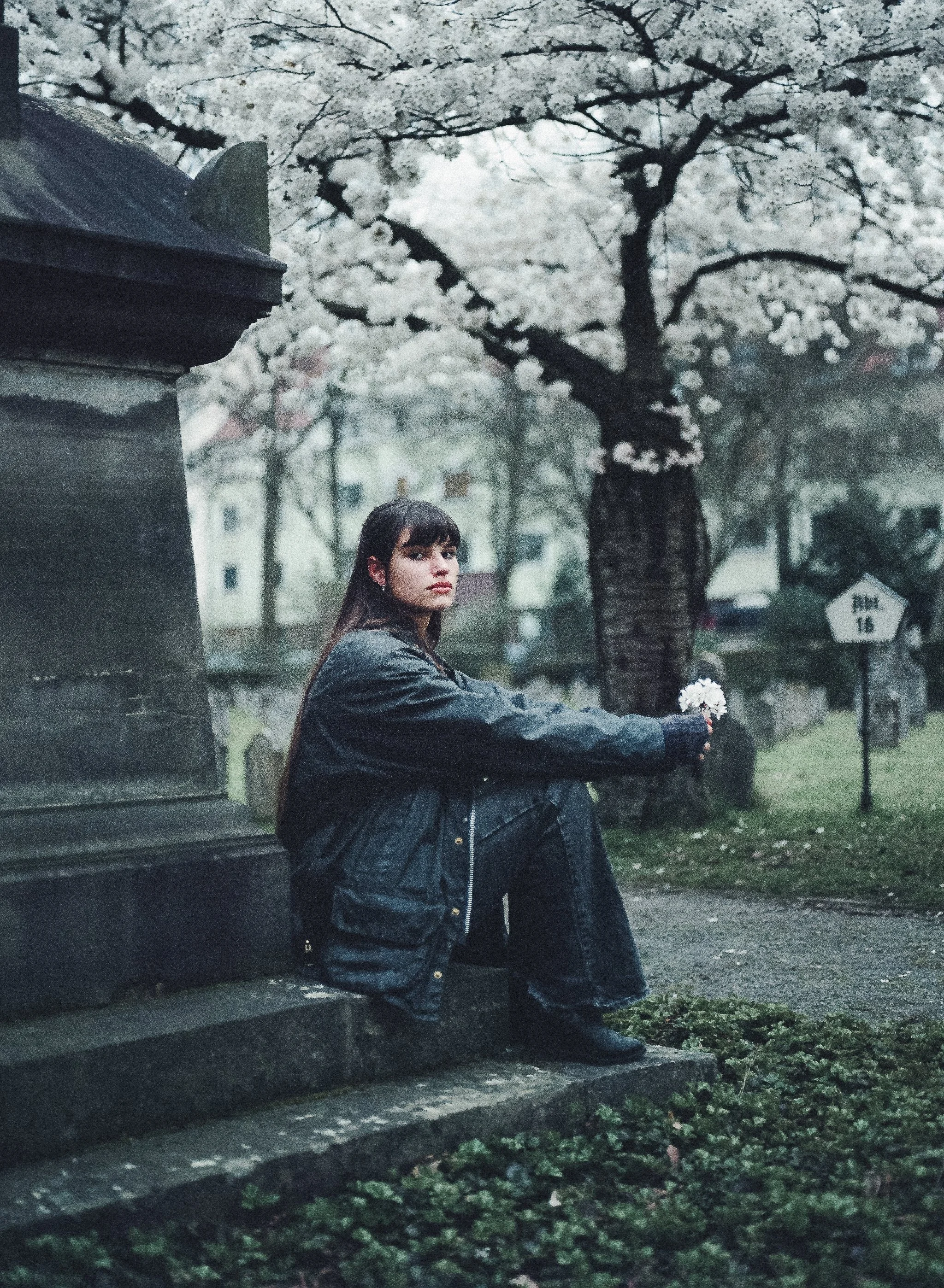 Eine junge Frau sitzt auf den Stufen eines Denkmals in einem Park, hält eine kleine Blumenstrauß, im Hintergrund blühende Kirschbäume, es ist gedämpftes Licht, typisch für einen Regentag.